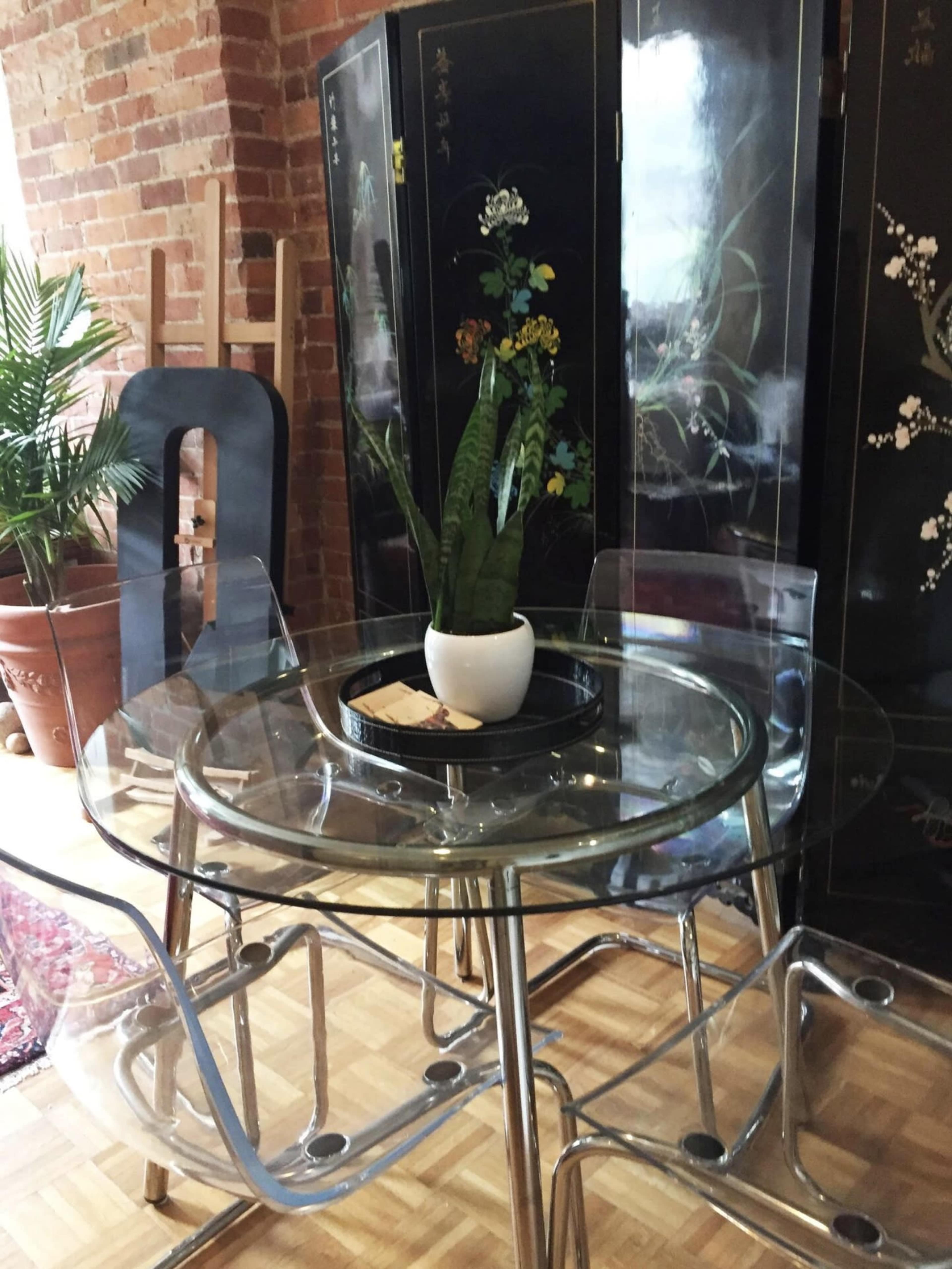 A glass-top dining table with clear chairs and a potted plant in the center, set against a backdrop of a brick wall and a decorative screen.