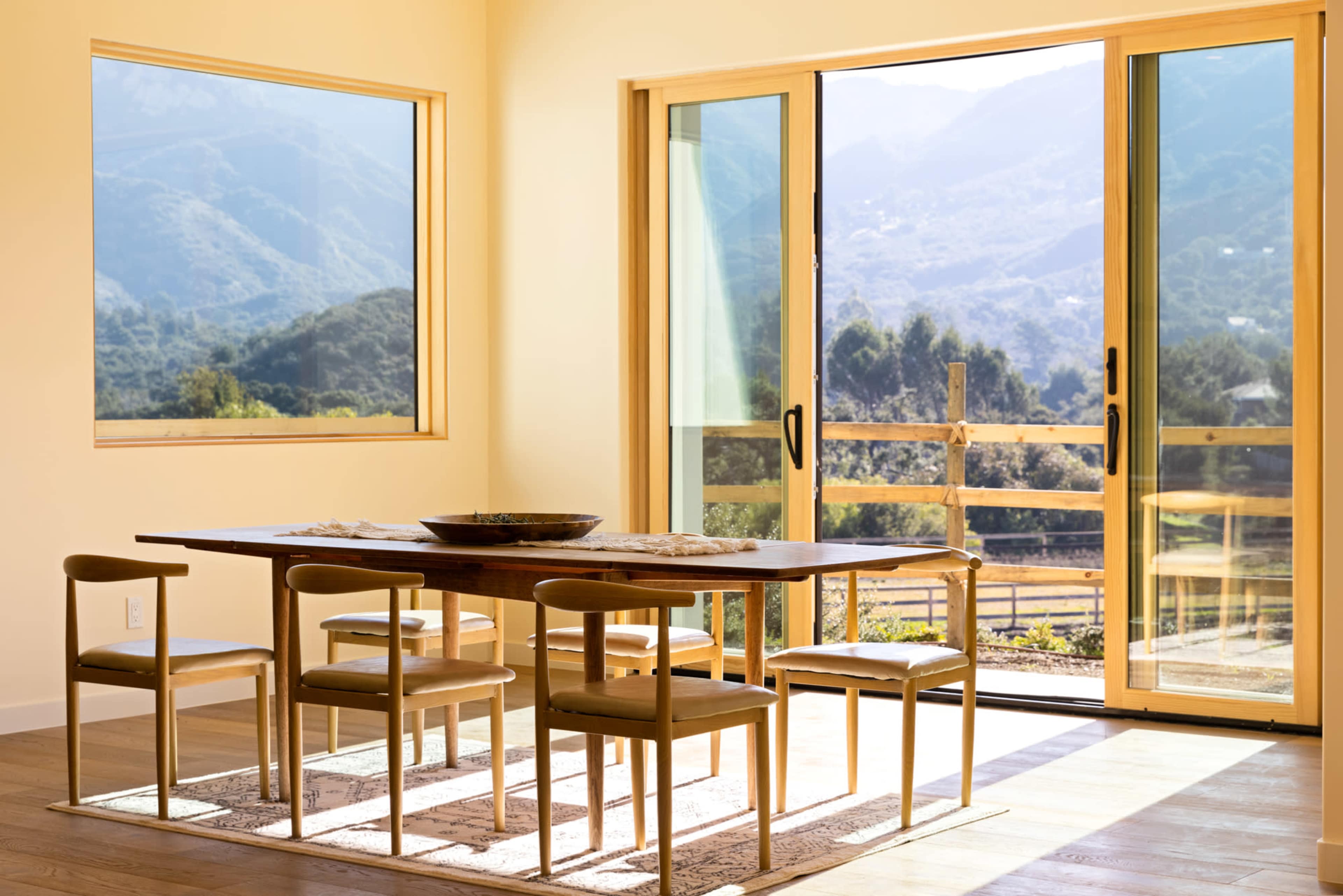 A wooden dining table with four chairs is positioned near a large sliding door that opens to a scenic view of mountains and greenery.