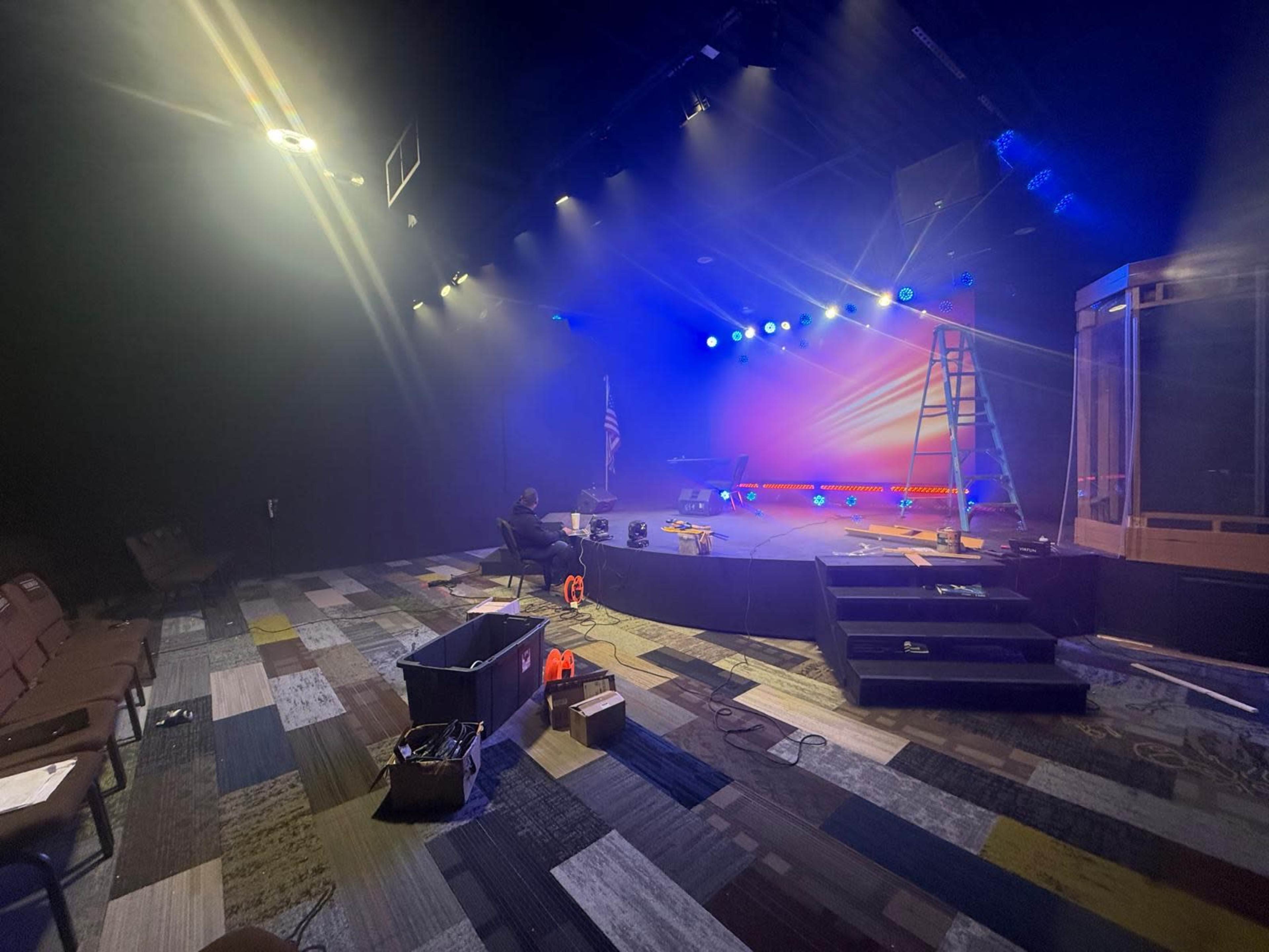 A stage is set up with a ladder, colorful lighting, and tools scattered around, indicating preparations for a performance or event.