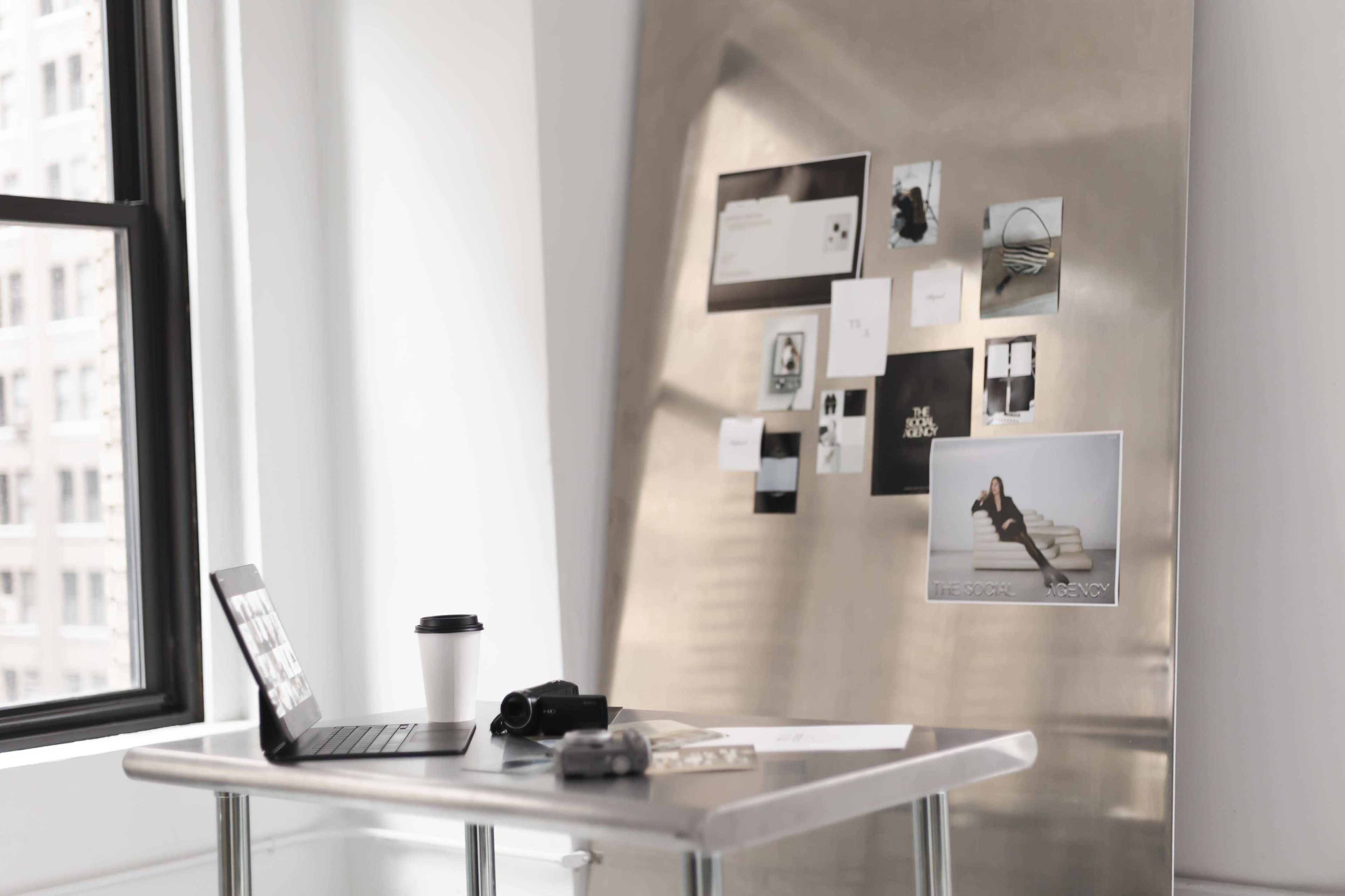 A sleek, modern workspace features a laptop on a table, a coffee cup, a camera, and a wall covered with various photos and notes.