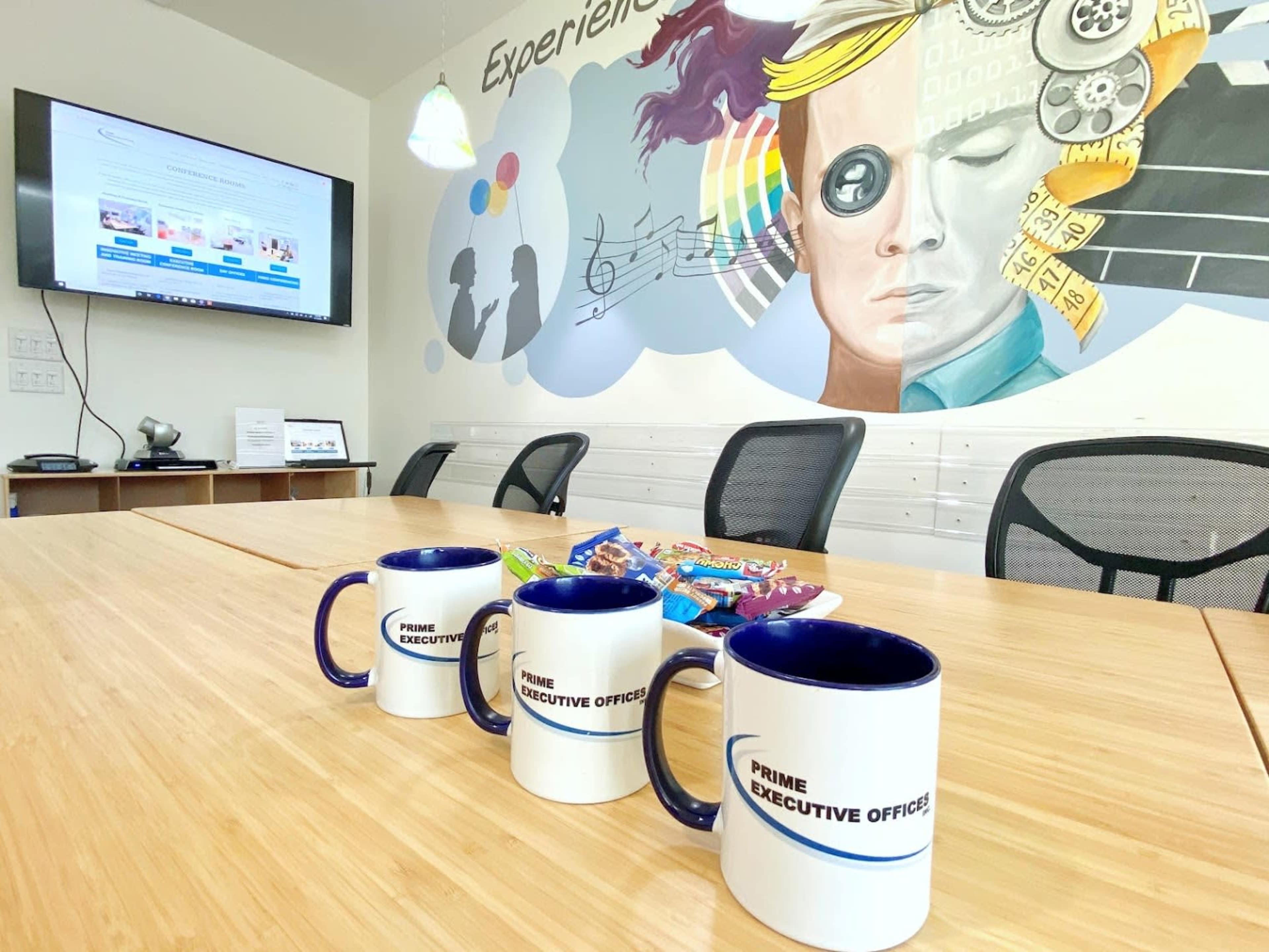 The image shows a conference room with a wall mural of a person's face, a television displaying a webpage, and three mugs arranged on a table alongside a plate of snacks.