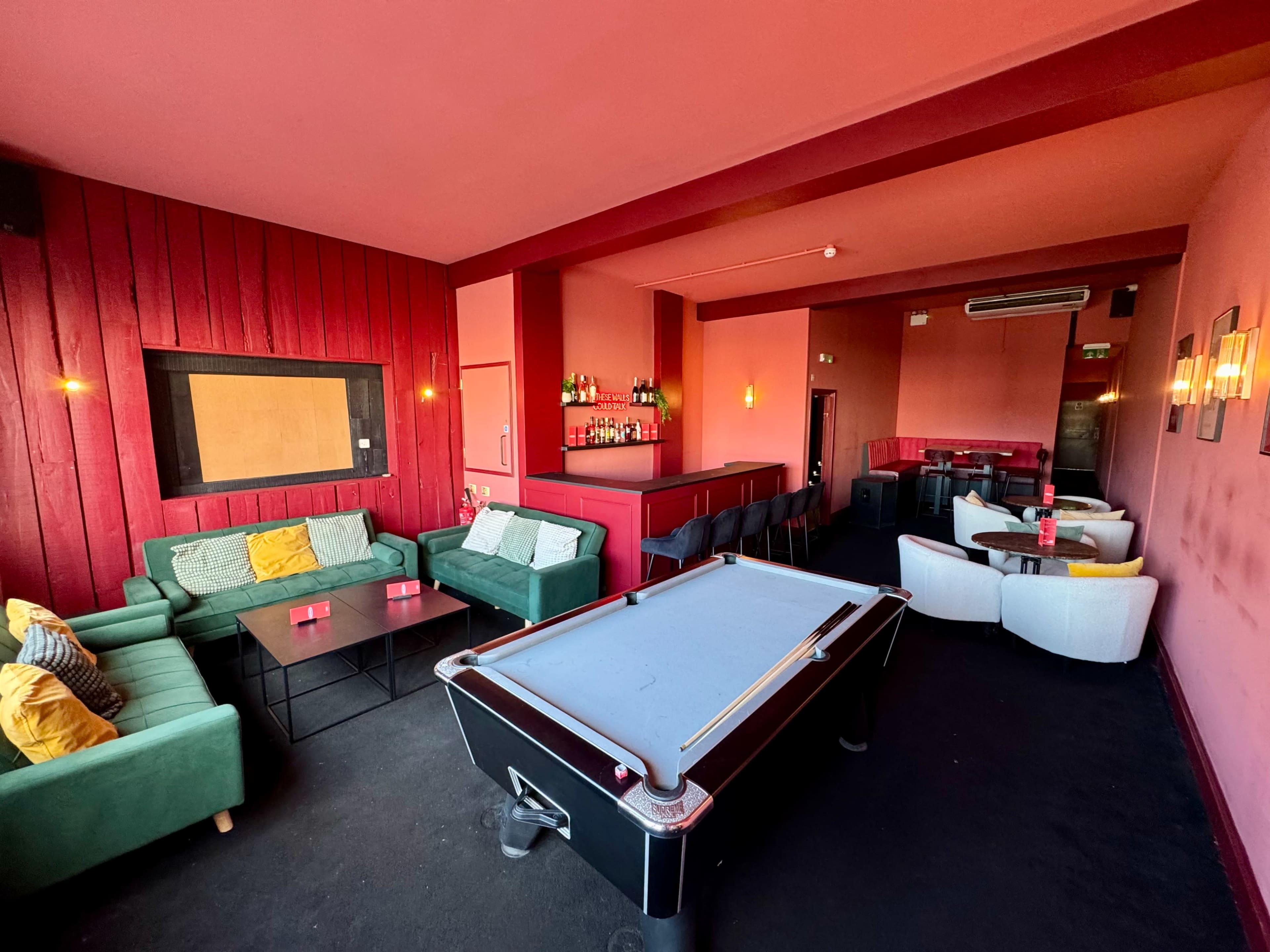 The image shows a stylish lounge area featuring a pool table, green sofas, a bar counter, and soft lighting in a room with pink walls.