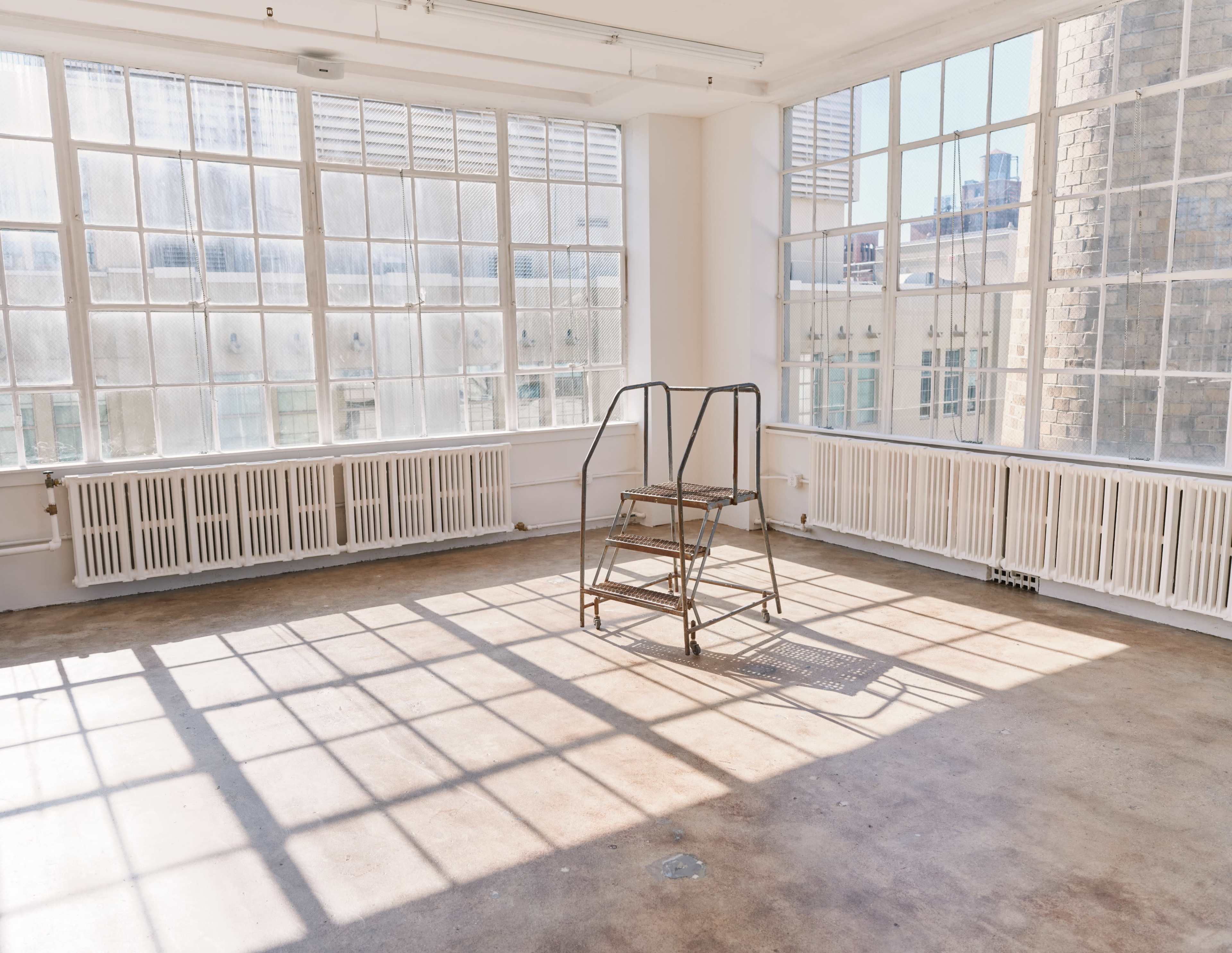 A metal step ladder stands in the center of a brightly lit, empty room with large windows and concrete flooring.