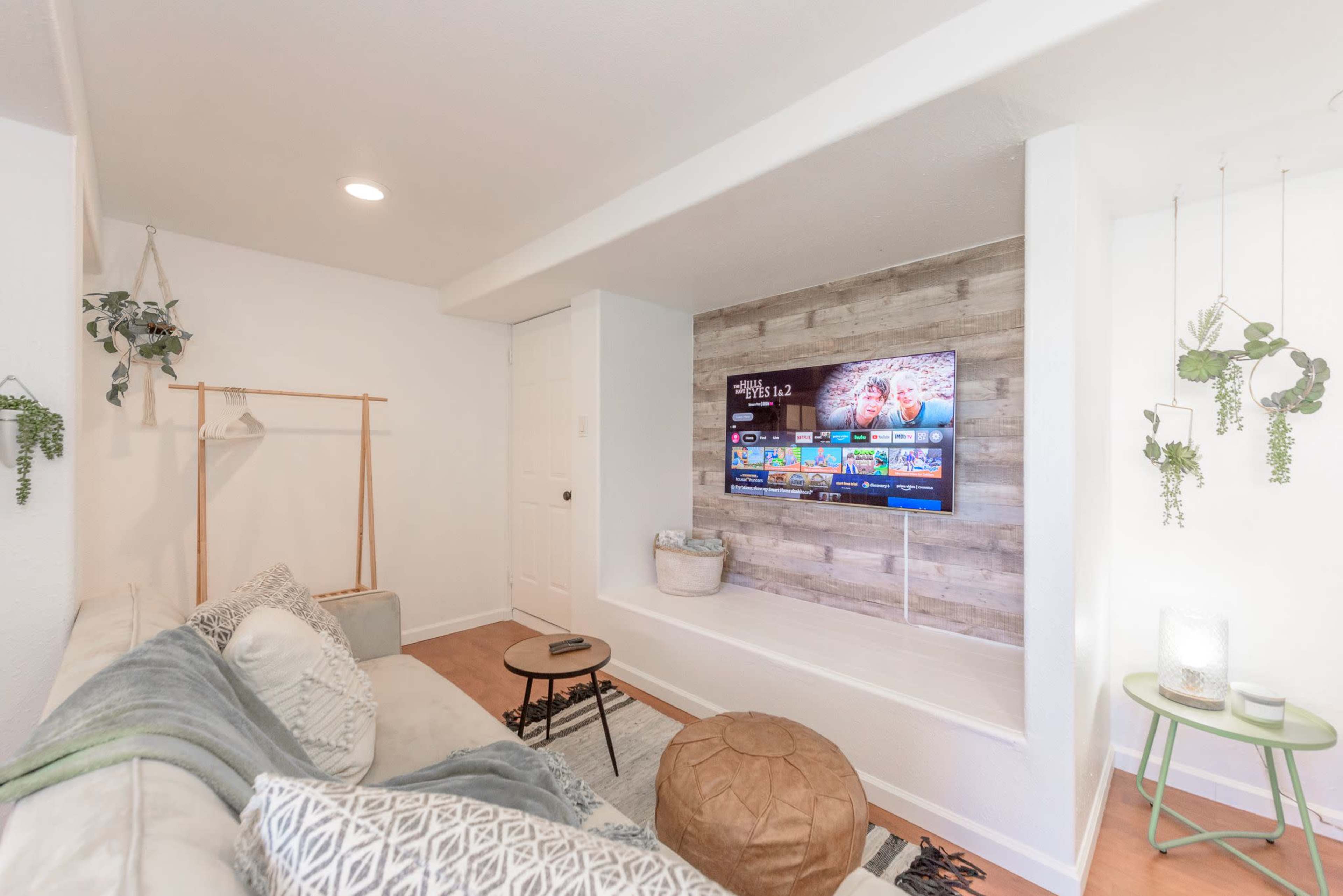 A modern living room with a wall-mounted TV and a sofa, featuring indoor plants and a light, minimalist decor.