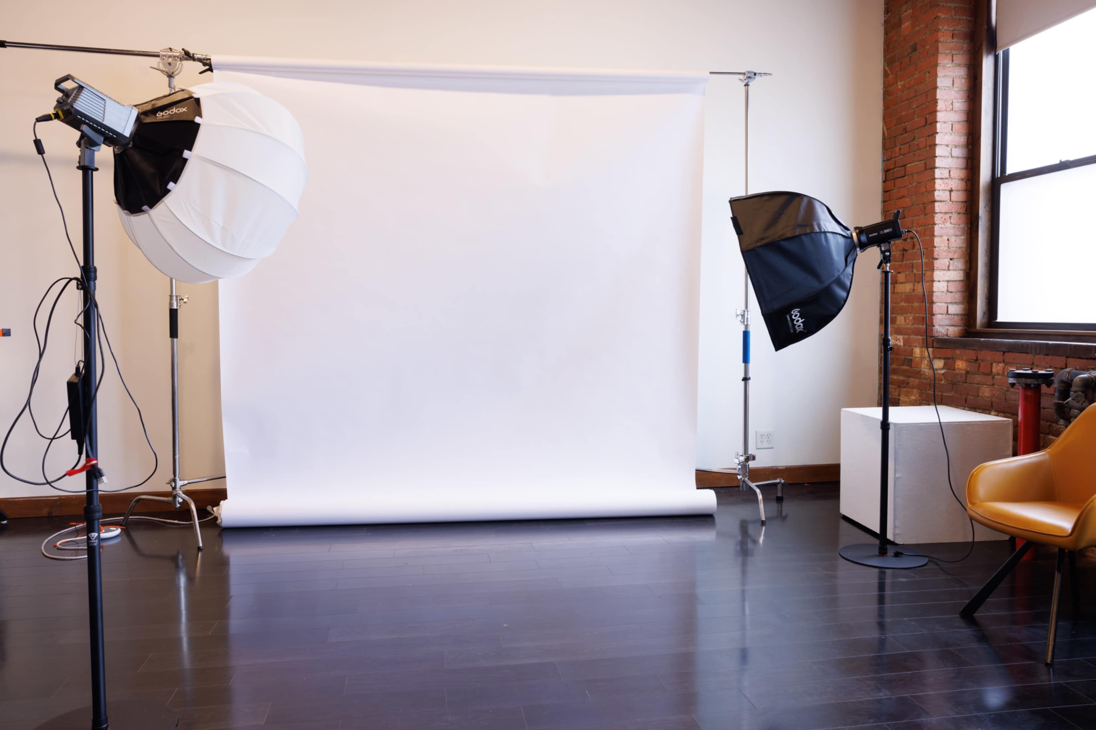 The image shows a photography studio with a white backdrop, studio lights on stands, and a chair set against a brick wall.