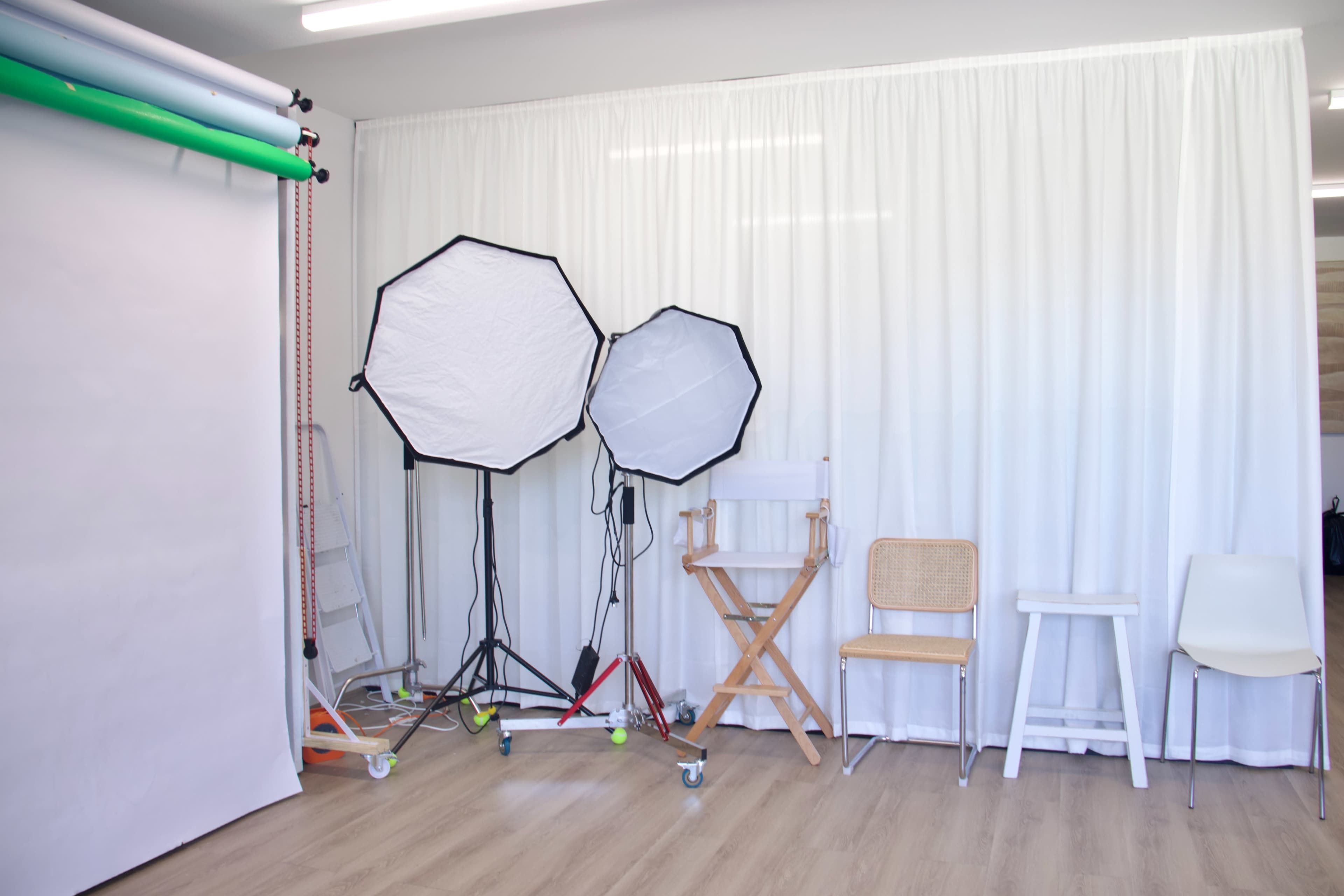 The image shows a photography studio setup with two softbox lights, a blank backdrop, and various seating options against a white curtain.