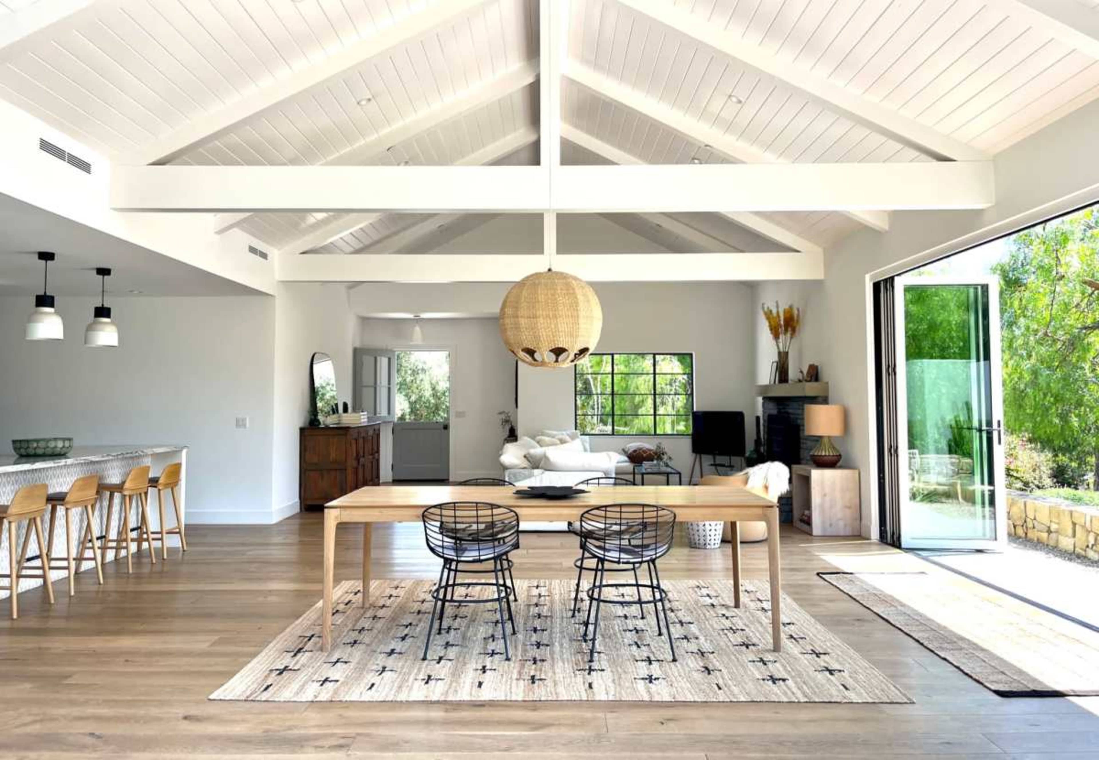 The image shows a spacious, modern living and dining area featuring a wooden table with black chairs, a large pendant light, and large glass doors that open to an outdoor space.