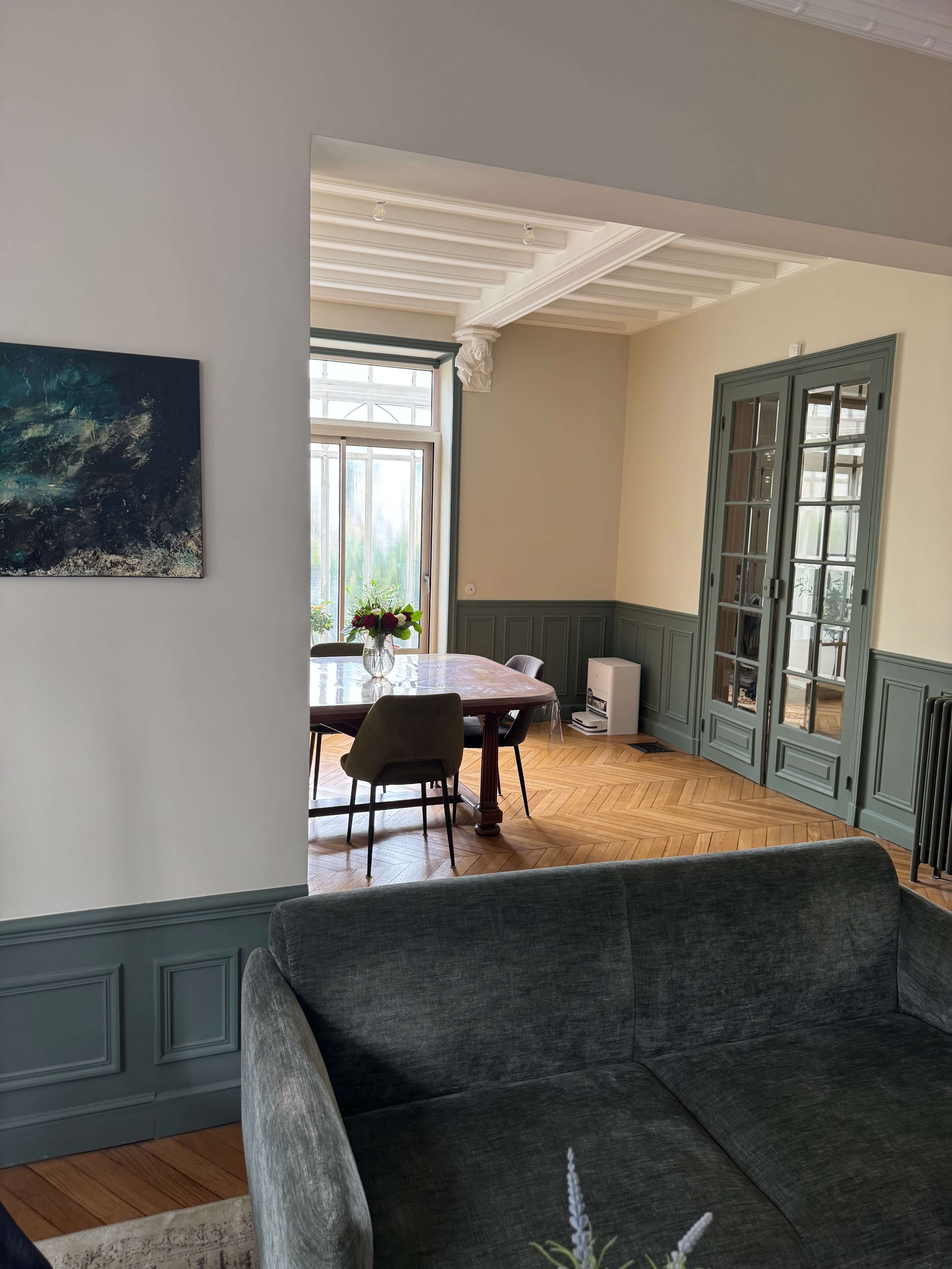 A dining area with a wooden table and chairs is visible through an archway, featuring large windows that allow natural light to enter the room.