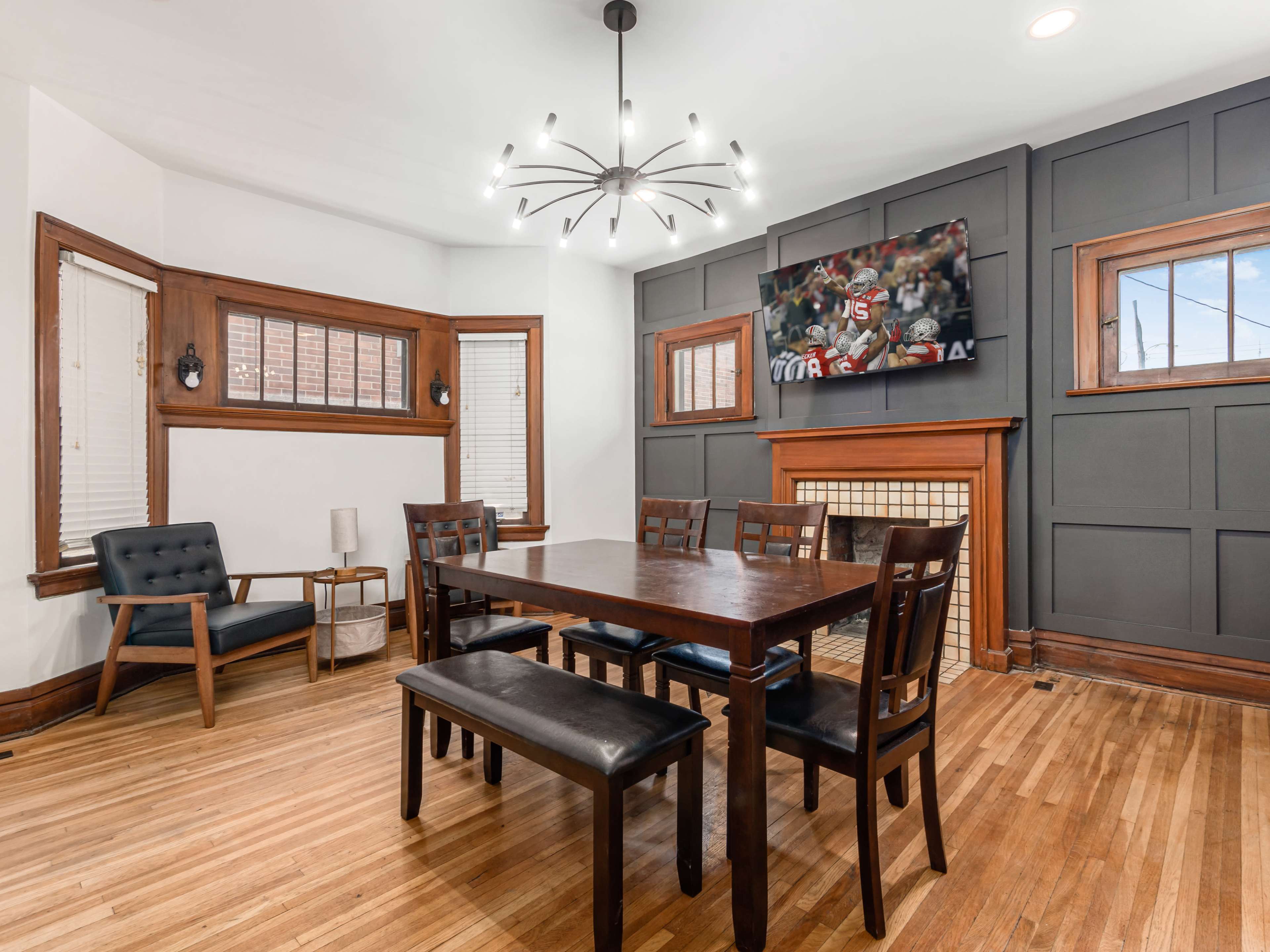 A dining area with a wooden table and chairs, a modern light fixture, and a television mounted on the wall, all set against paneled walls and hardwood flooring.
