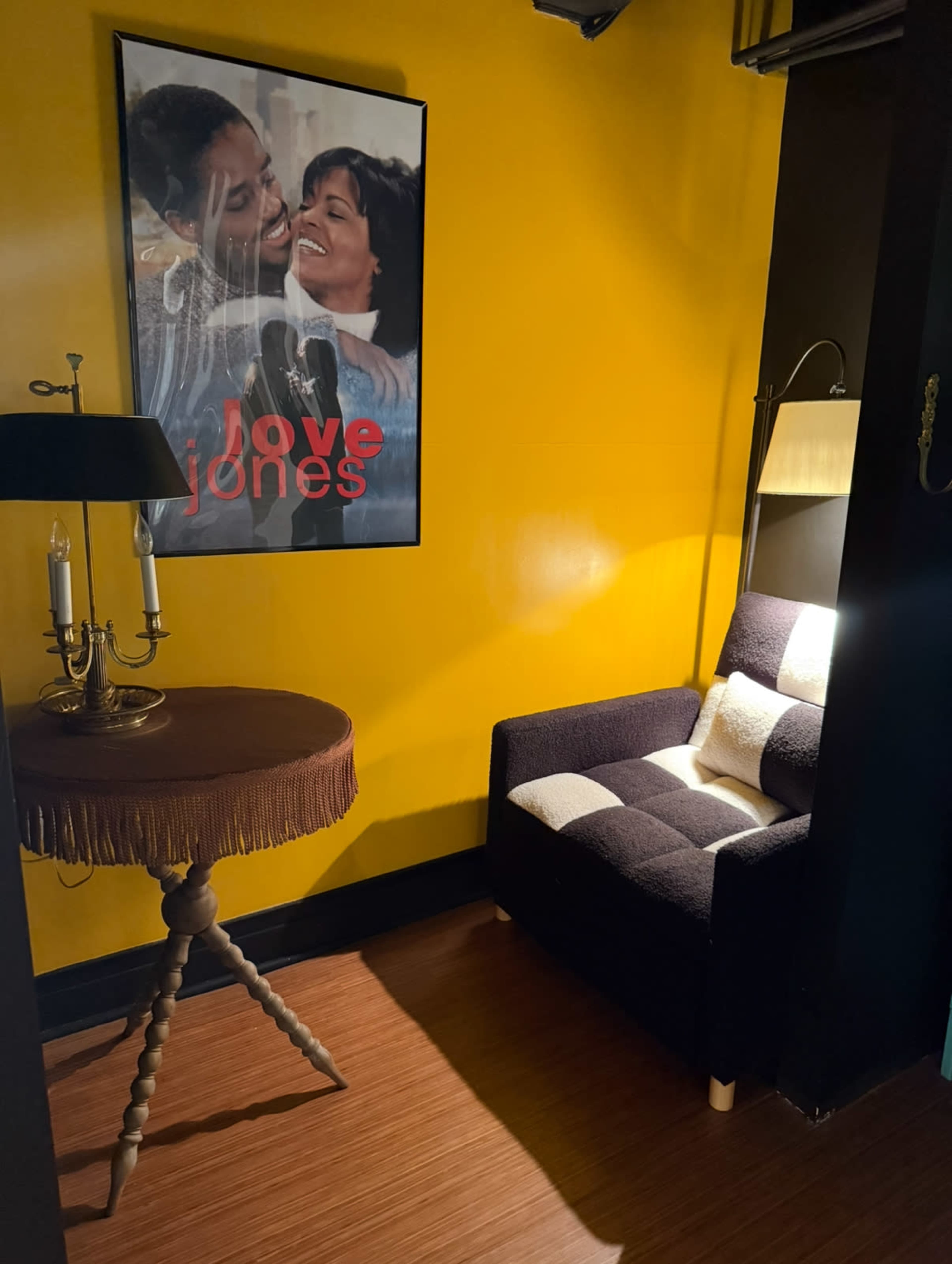 A cozy corner features a black and white upholstered chair beside a round table with a fringed edge, illuminated by a lamp, and a vibrant yellow wall adorned with a "Love Jones" movie poster.
