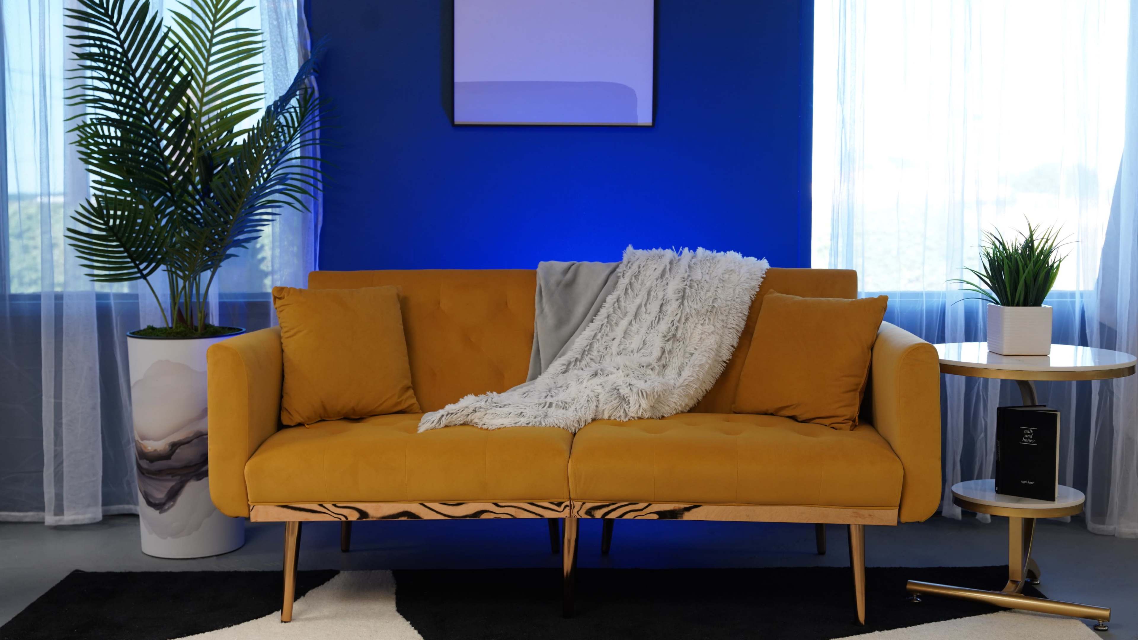 A mustard yellow sofa with a white throw blanket is positioned against a blue wall, accompanied by a plant and a small side table.