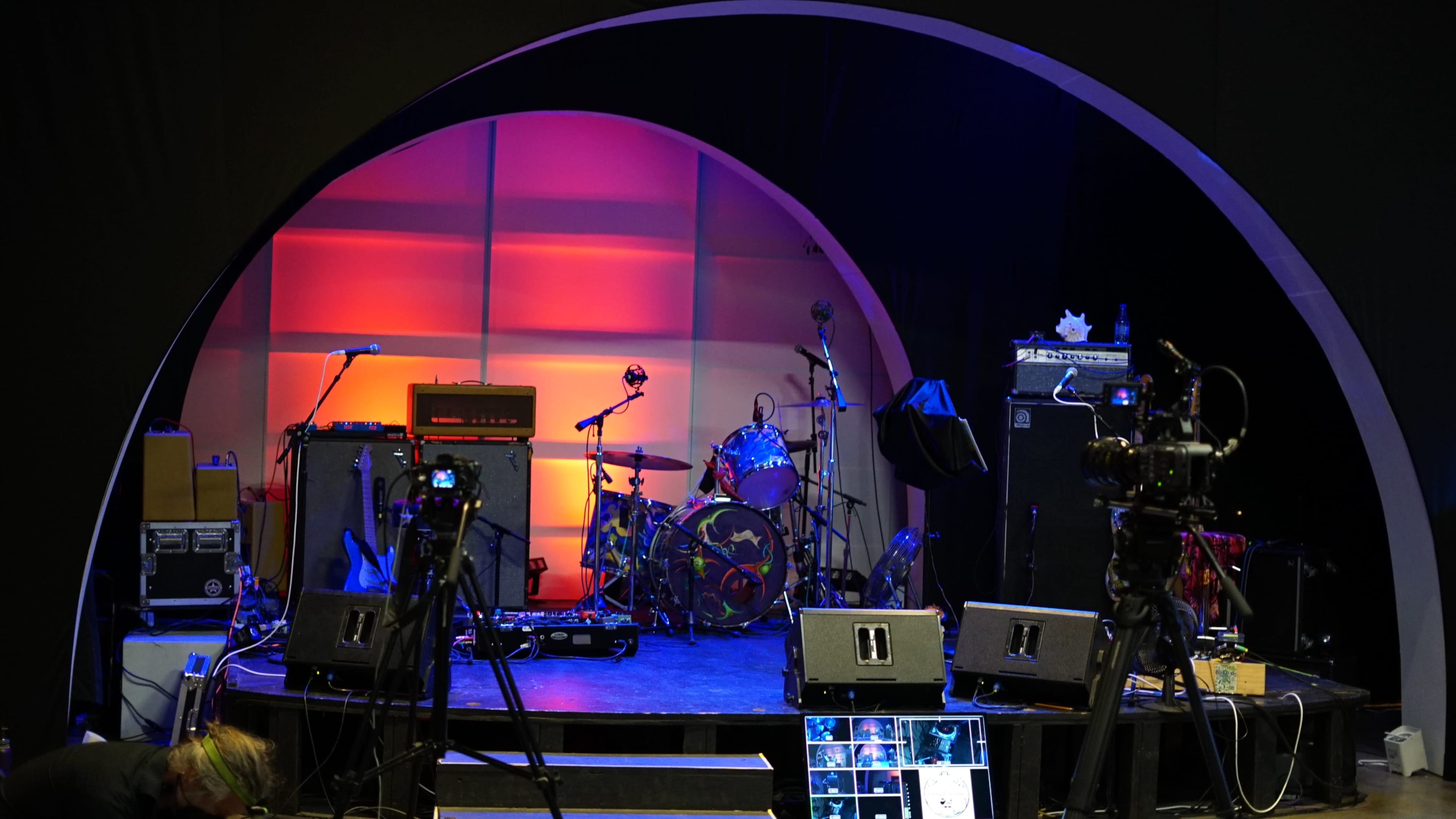 A stage setup with musical instruments, amplifiers, and cameras arranged in a semi-circular backdrop.