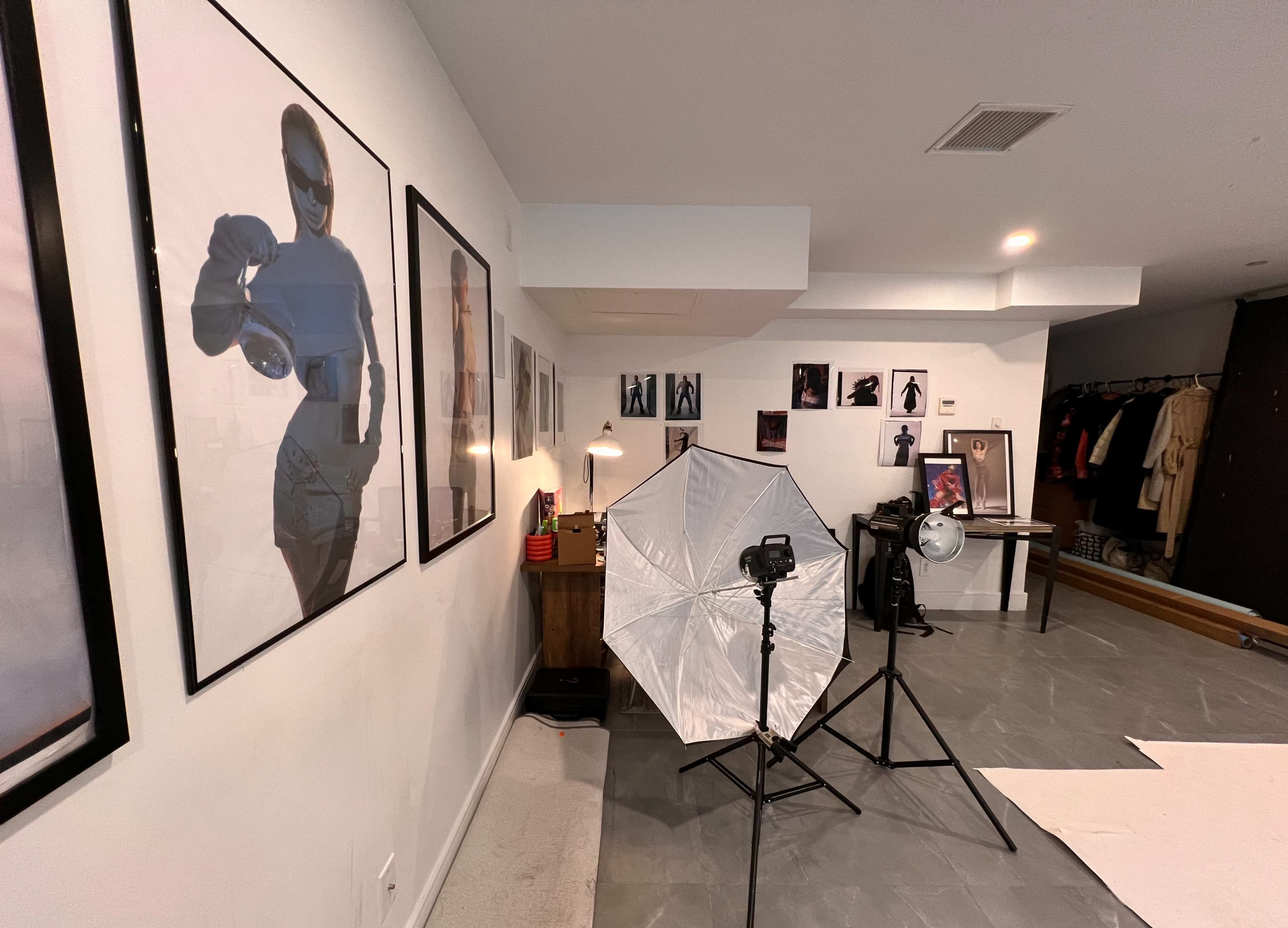 The image shows a photography studio featuring various framed photographs on the walls, a lighting setup with an umbrella, and a camera on a tripod facing a backdrop.