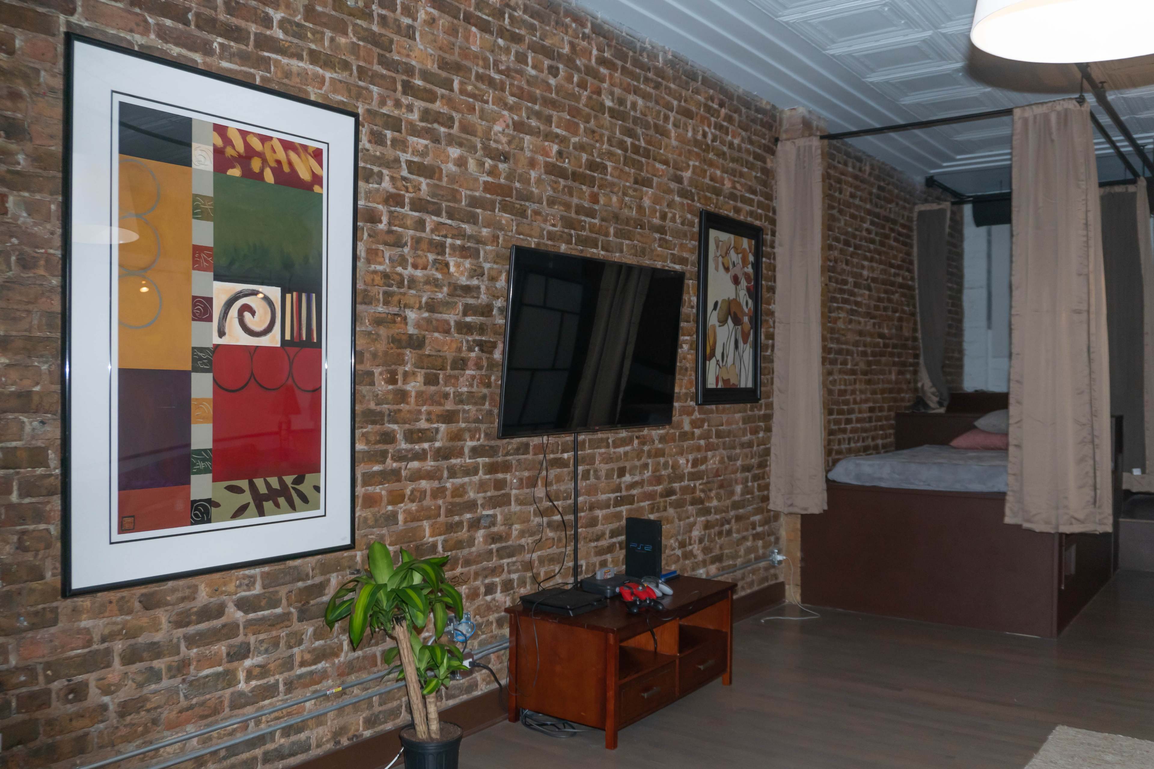 A room featuring exposed brick walls, framed artwork, a flat-screen TV, a wooden entertainment unit, and a bed partially enclosed by curtains.