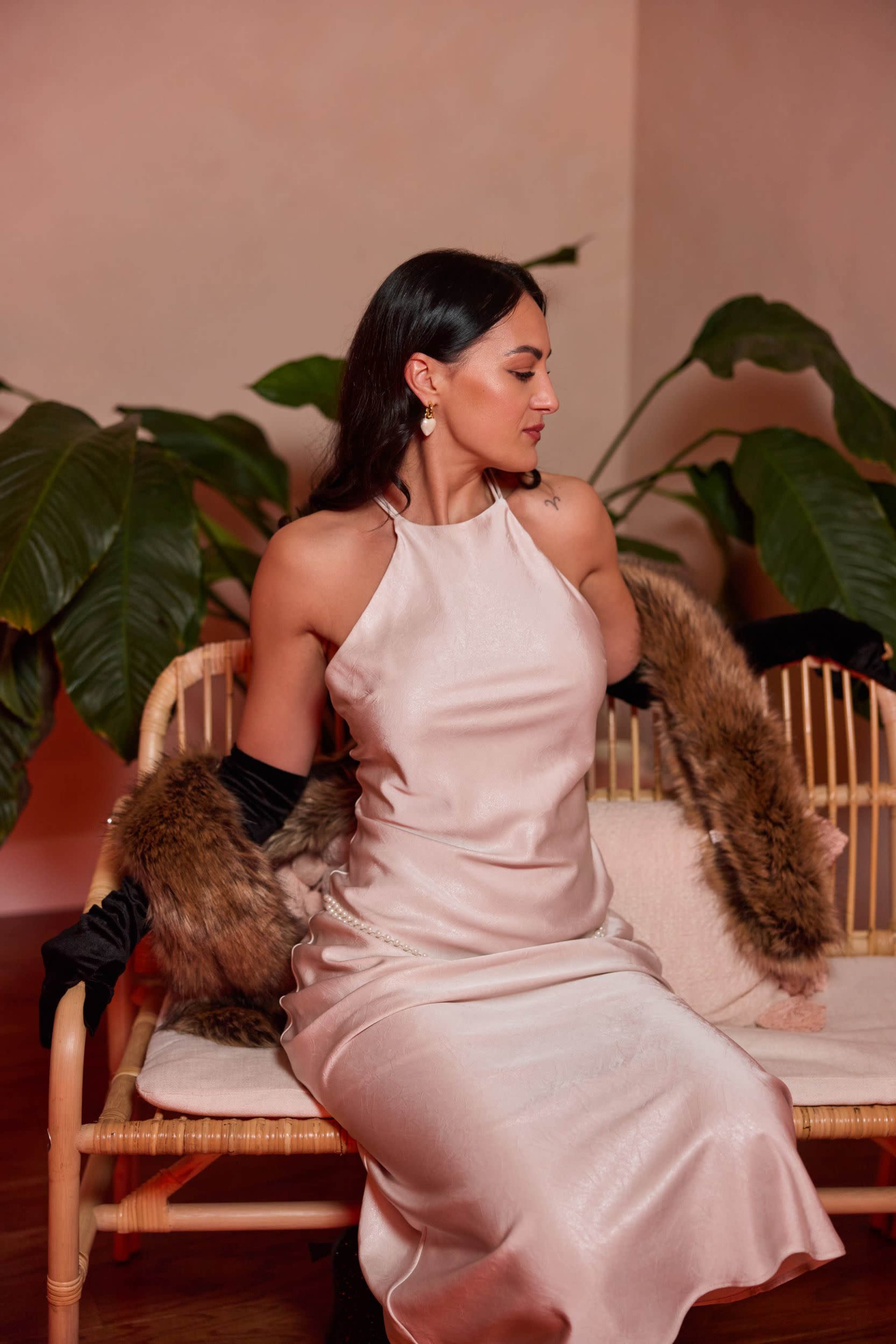 A woman in a satin dress sits on a rattan couch surrounded by large leafy plants.