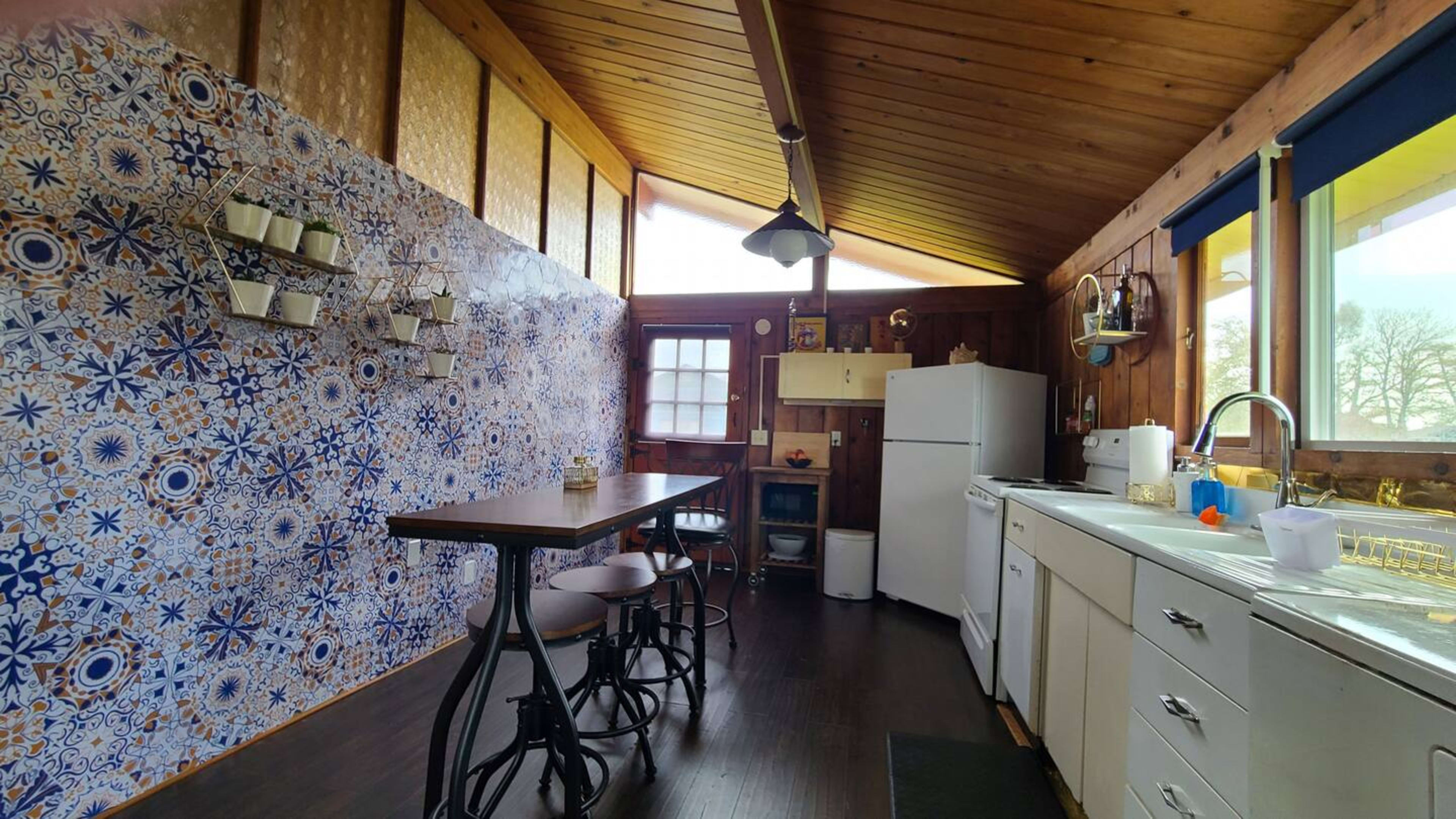 The image shows a kitchen with a tile accent wall, a wooden ceiling, a white fridge, a sink with a window, and a dining table with three stools.
