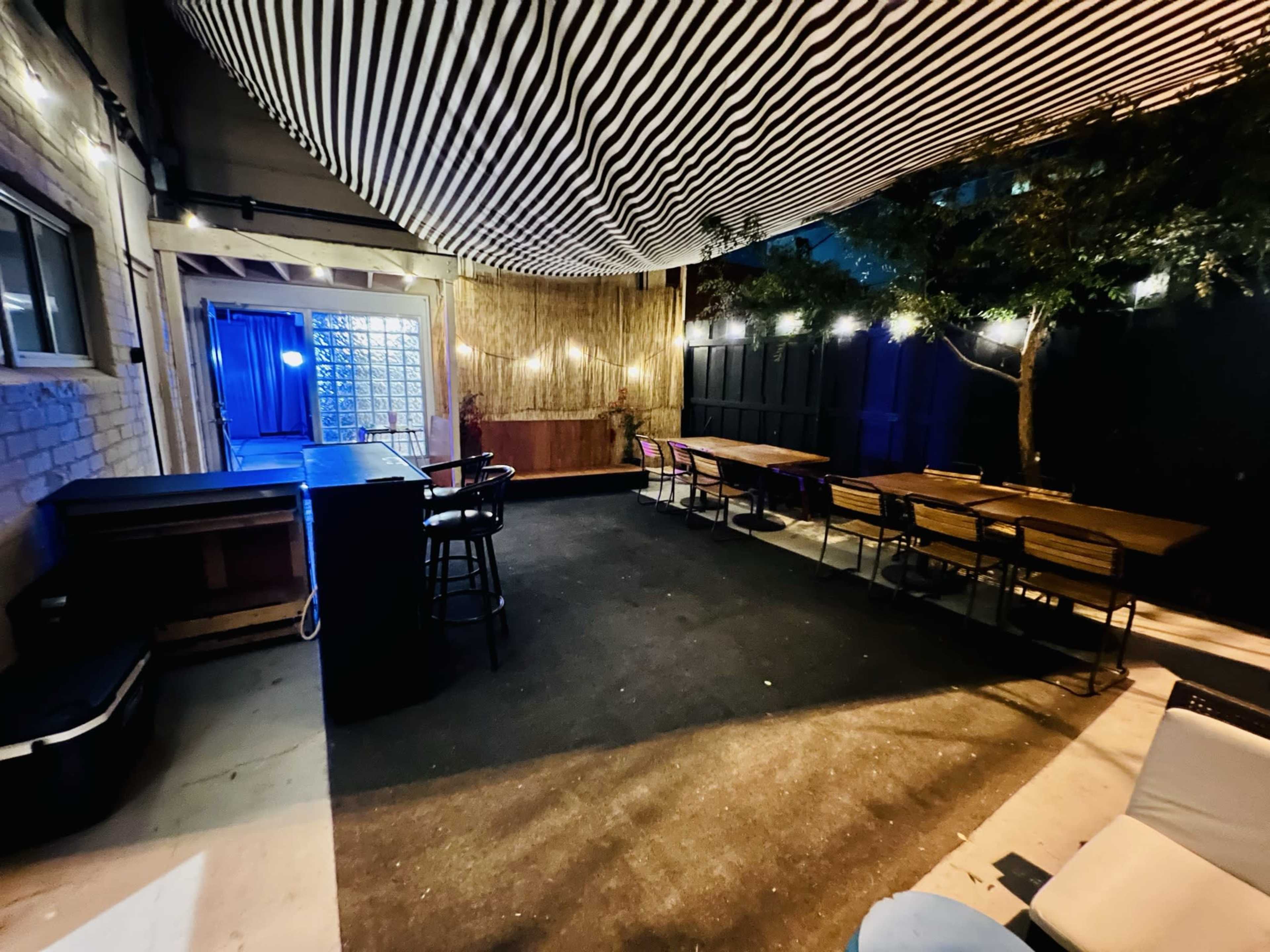 An outdoor patio area with a striped canopy, bar seating on one side, and several dining tables arranged on the opposite side, illuminated by string lights.