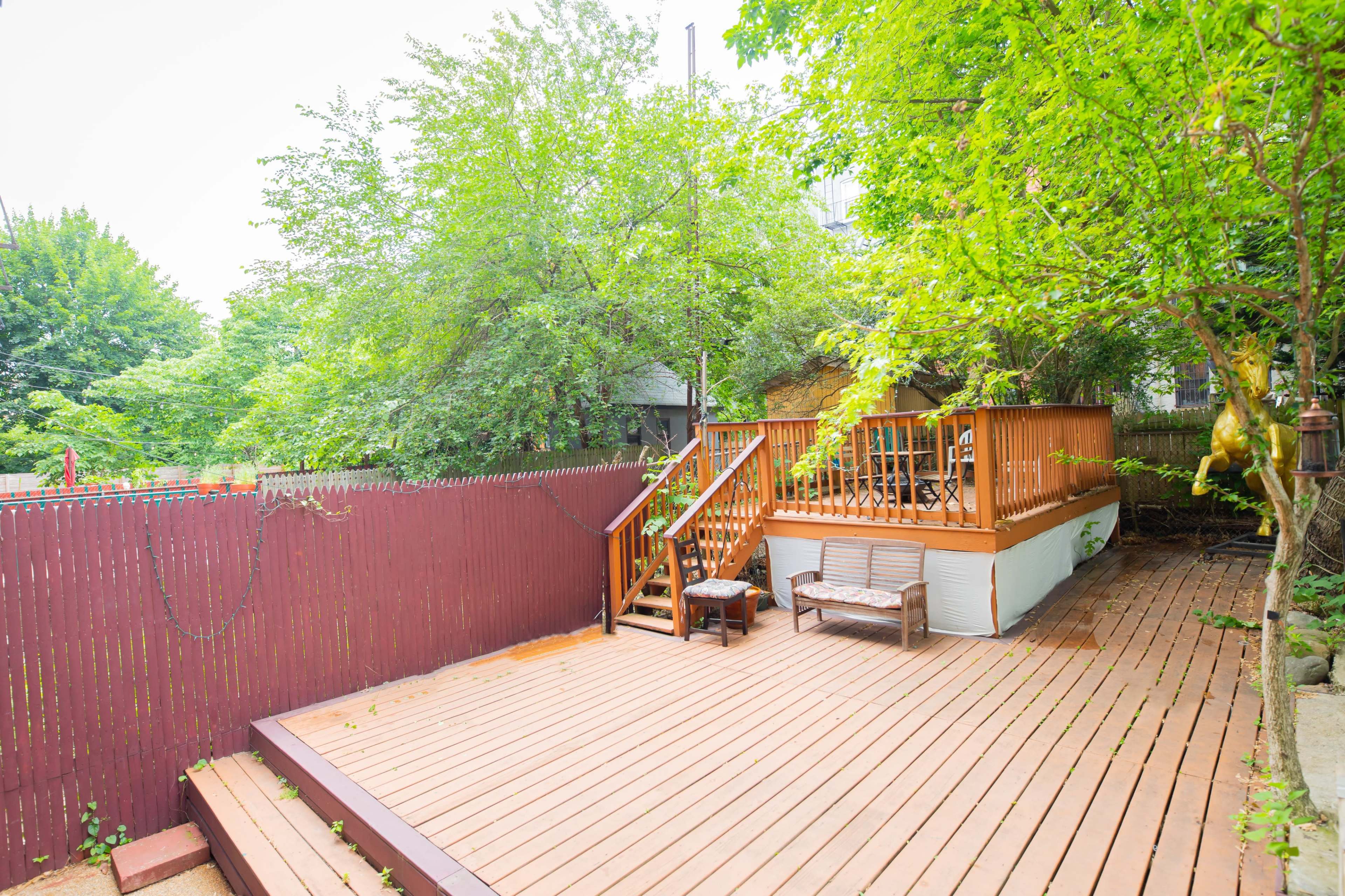 A wooden deck with a staircase leading to a raised area surrounded by greenery and a red fence.