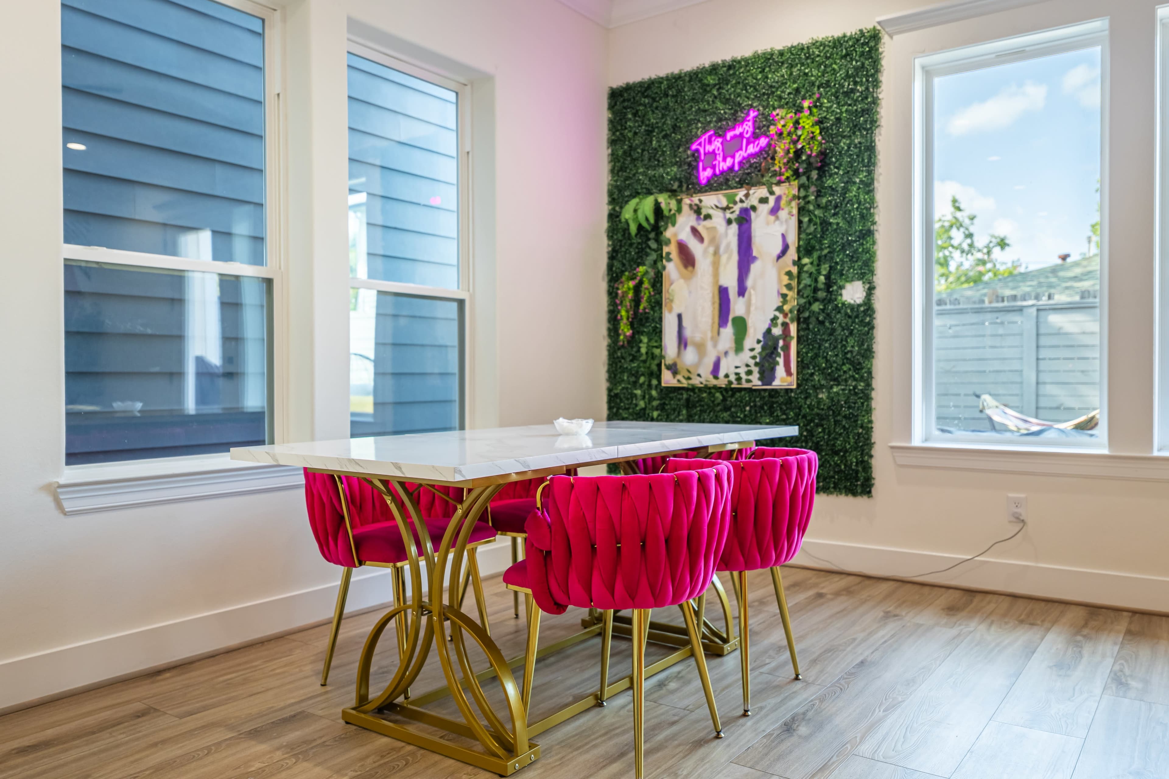 The image shows a dining area featuring a marble table and three pink tufted chairs, with a green wall decoration and a neon sign.