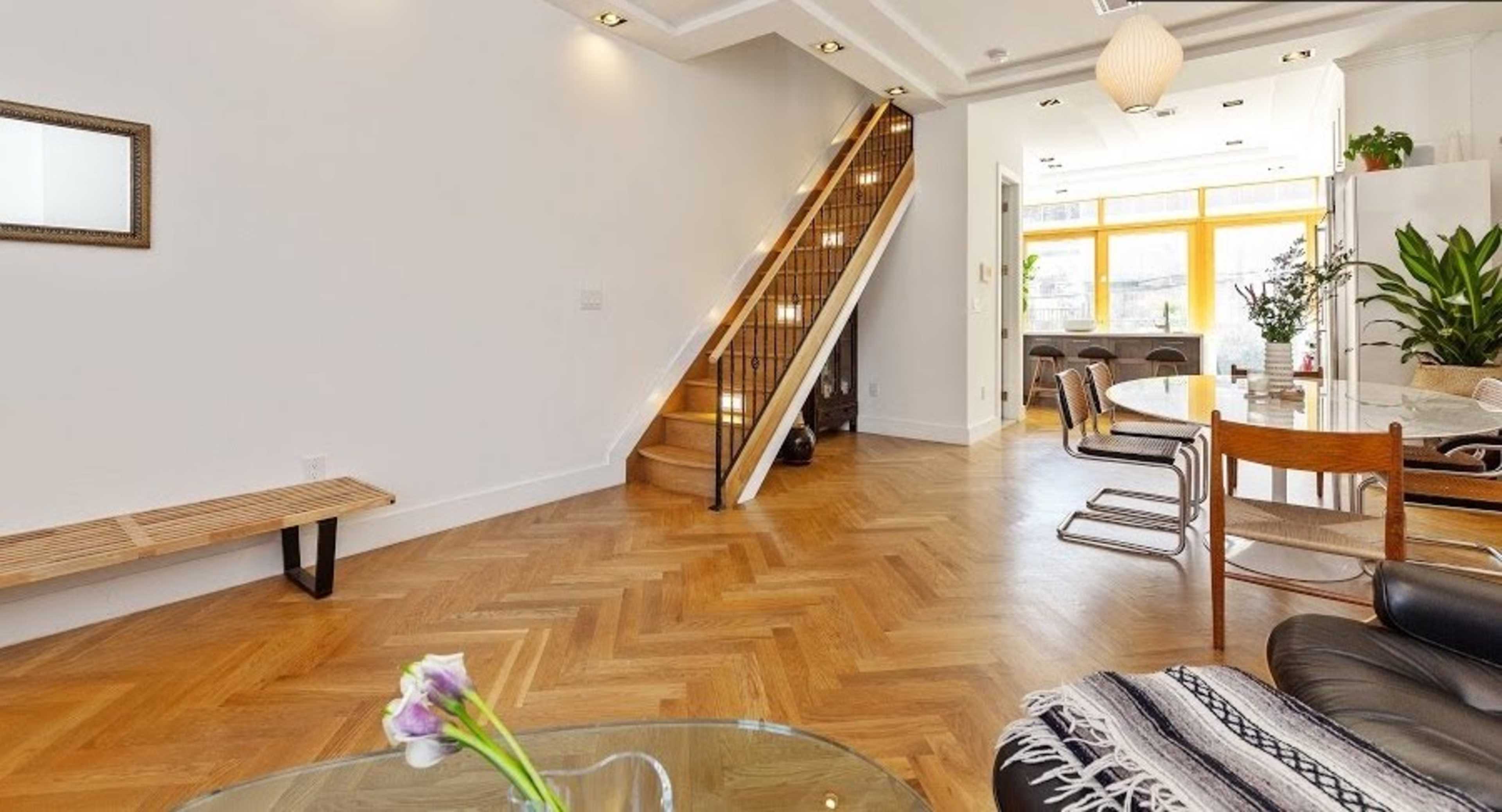 A spacious interior room featuring wooden herringbone flooring, a staircase along one wall, and a dining area with modern furniture.