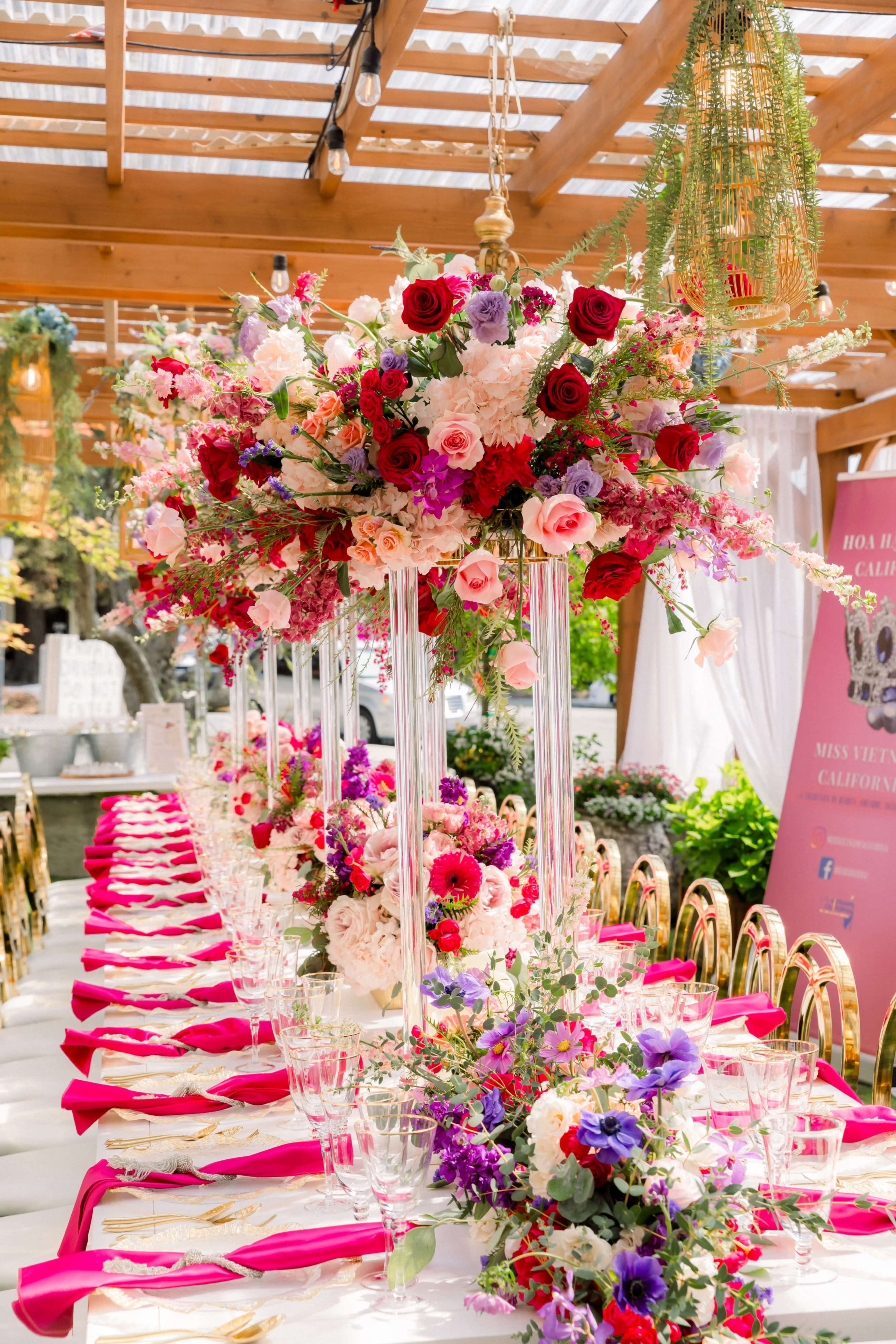 A long banquet table is adorned with vibrant floral centerpieces and elegant place settings, set under a wooden pergola.