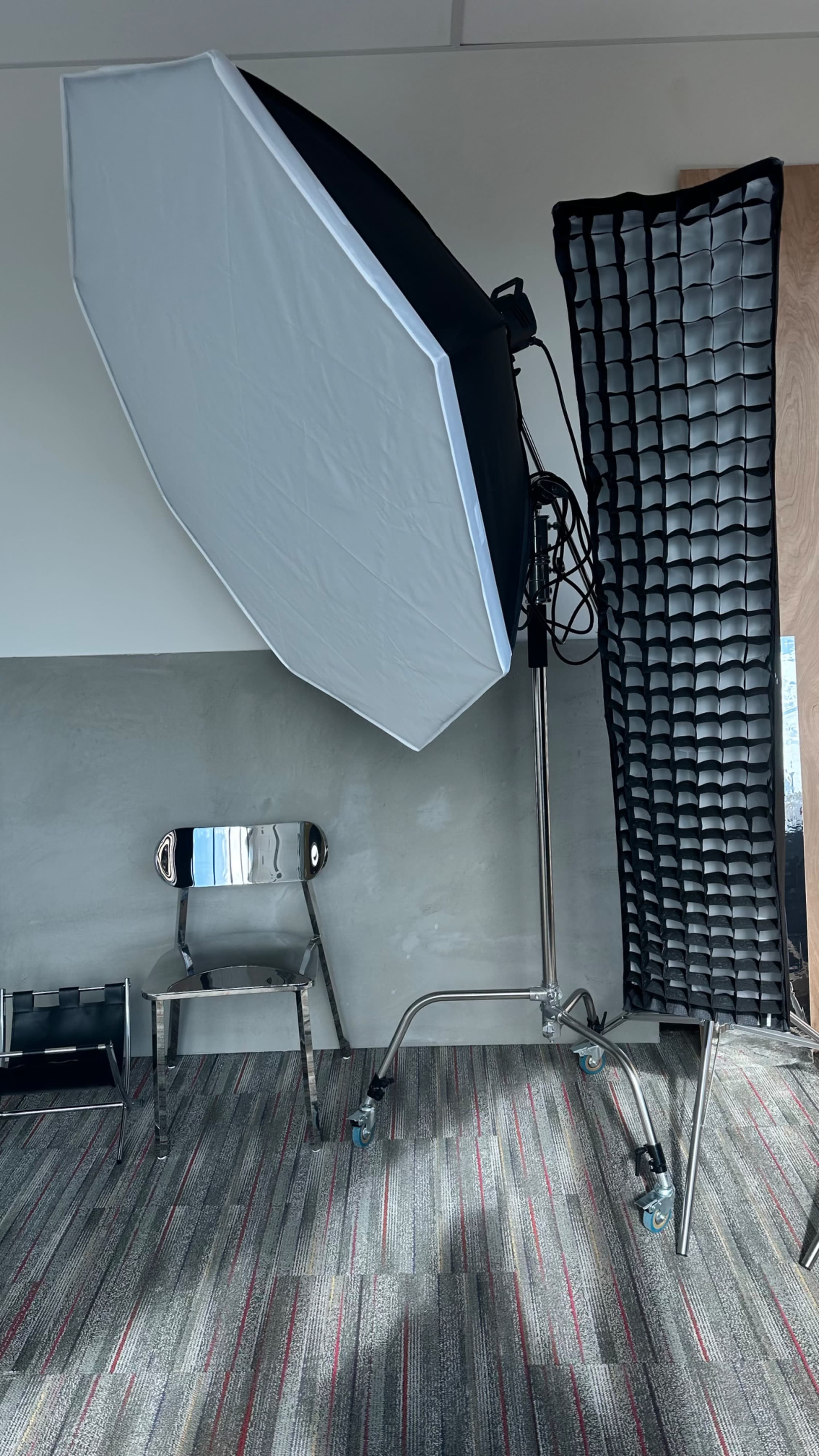 A photography setup featuring an octagonal softbox on a stand next to a folded, textured backdrop and a metal chair on a patterned carpet.