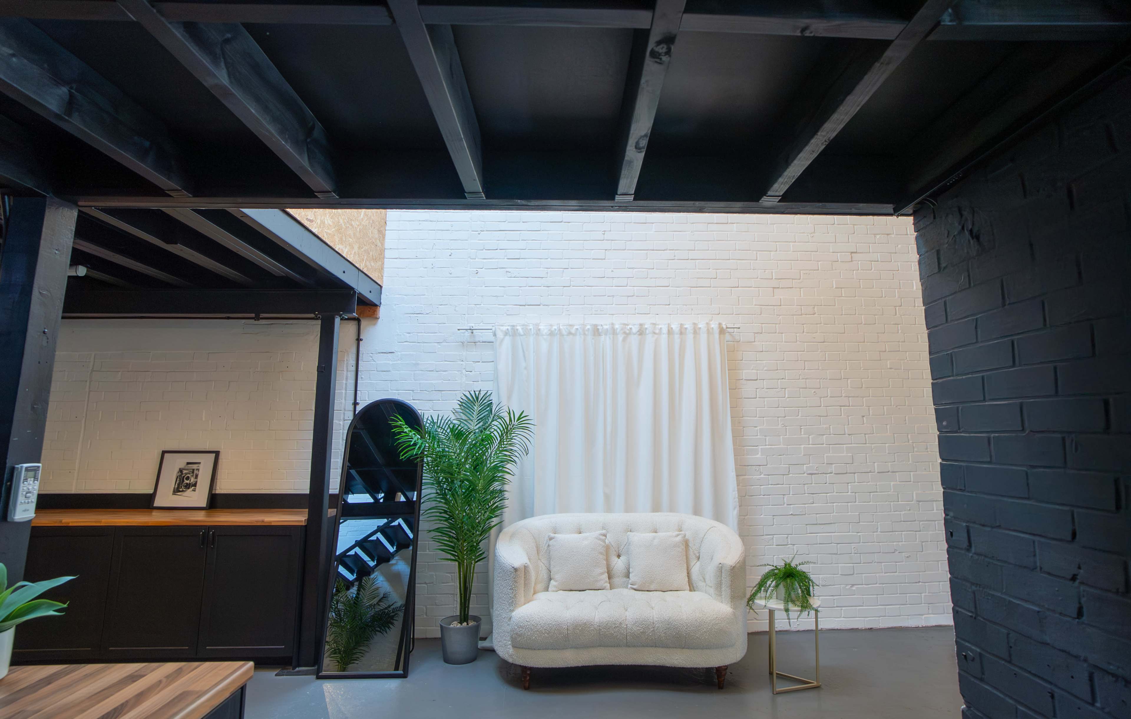A white sectional sofa is positioned in front of a curtained window in a modern, minimalistic interior space with exposed beams and potted plants.