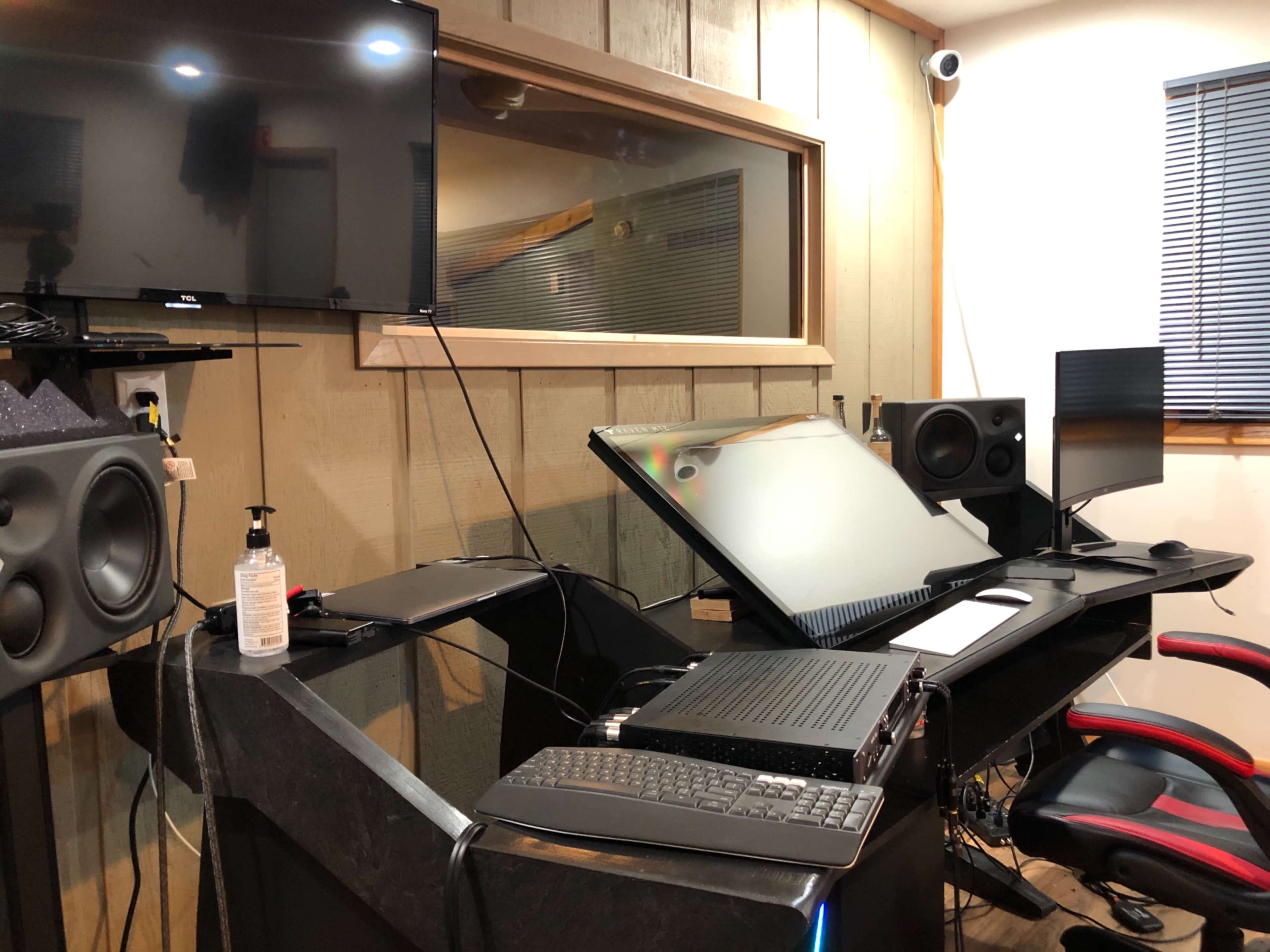 A music studio setup with a large mixing console, multiple monitors, speakers, and an ergonomic chair against a wooden-paneled wall.