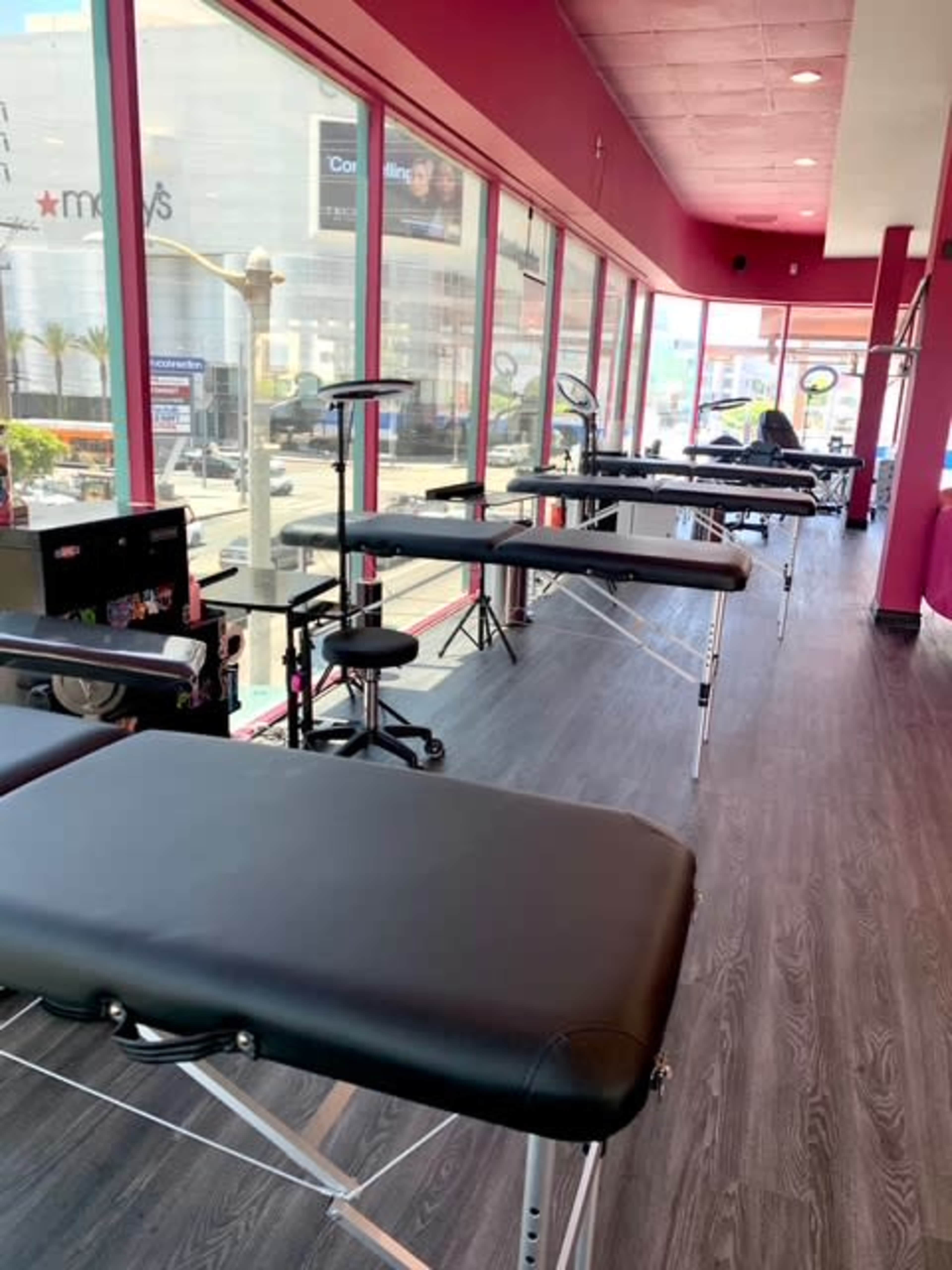 The image shows a brightly lit room with several black massage tables arranged in a line next to large windows.