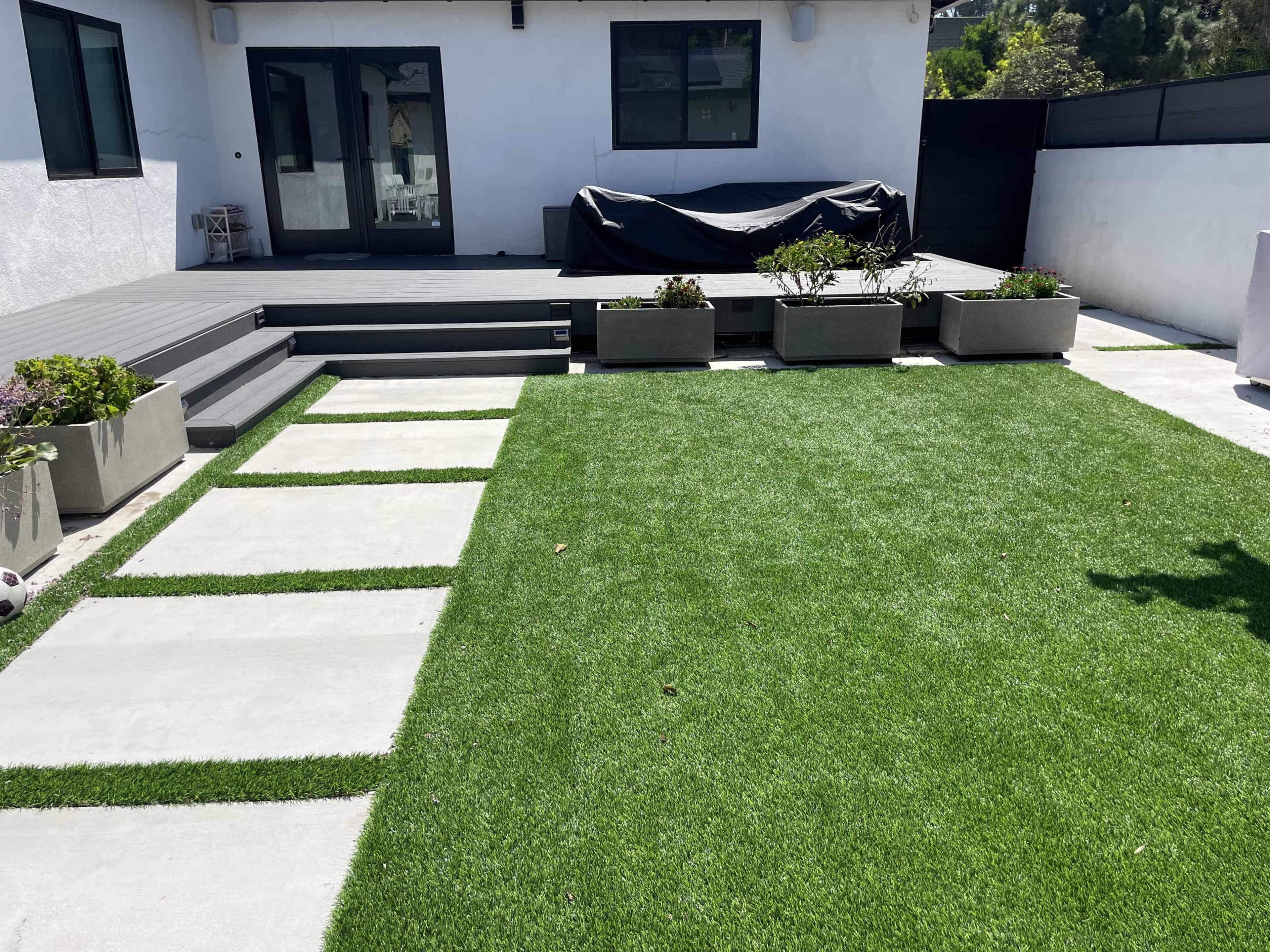 A backyard featuring artificial grass, concrete stepping stones, and a covered seating area with a large outdoor table.