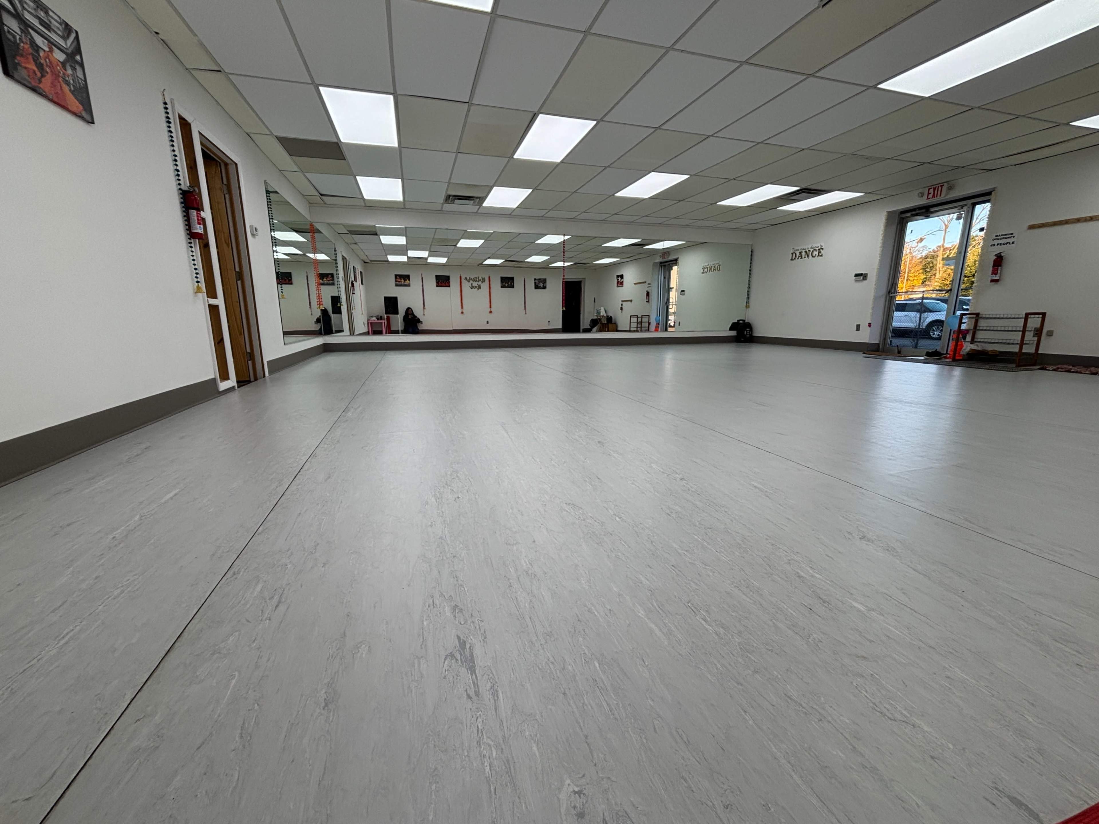 The image shows a spacious, empty room with smooth gray flooring and large windows allowing natural light to enter.