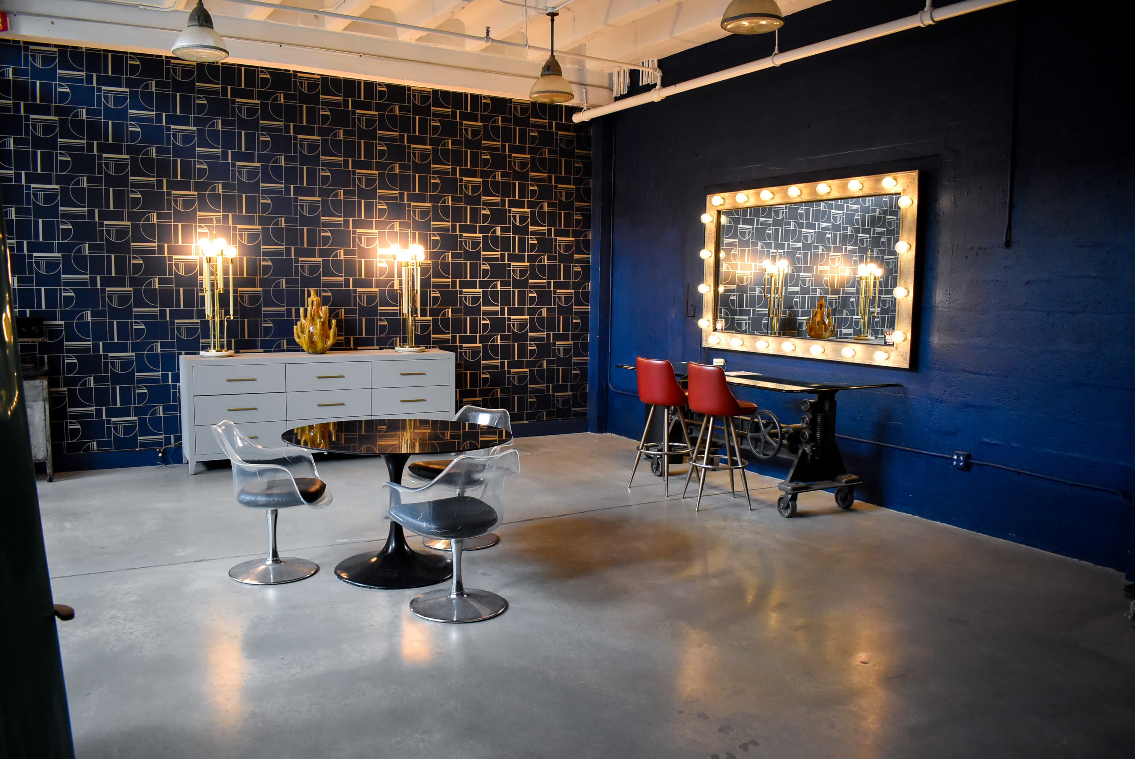 A spacious room with a dark patterned wall, modern furniture, a large illuminated mirror, and a table with red chairs.