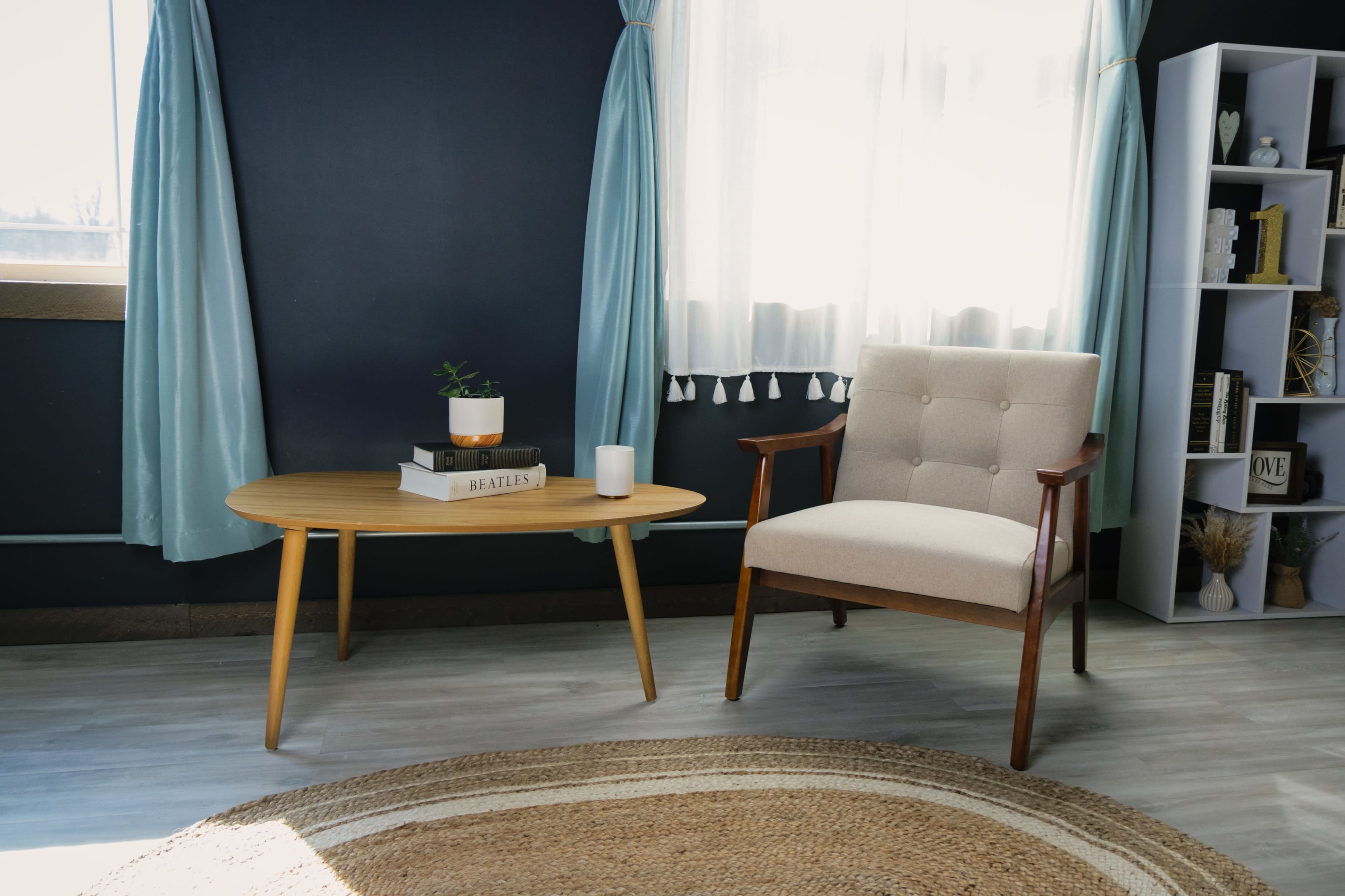 A modern living room features a light wooden table, a beige upholstered chair, and a woven rug on a floor with large windows dressed in pale blue curtains.