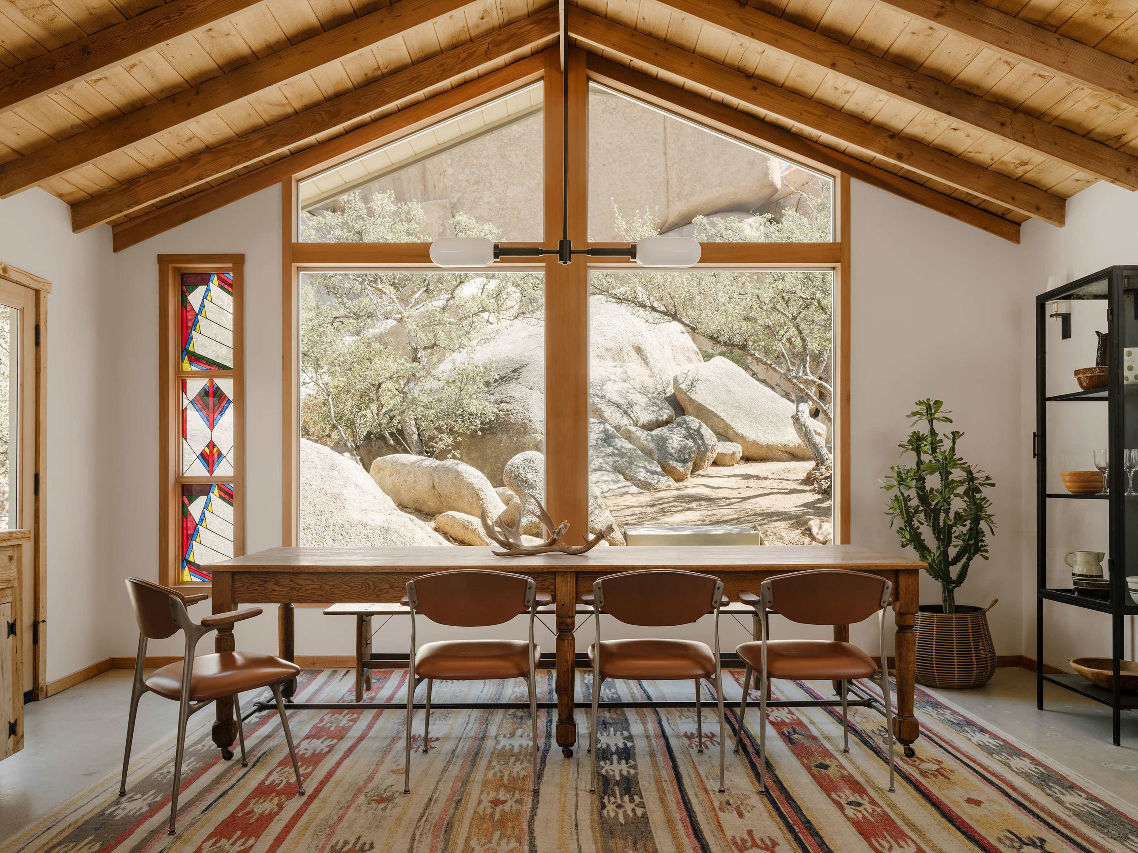 A spacious dining area with a wooden table and chairs, large windows that reveal a rocky outdoor landscape, and a vaulted ceiling with exposed wooden beams.