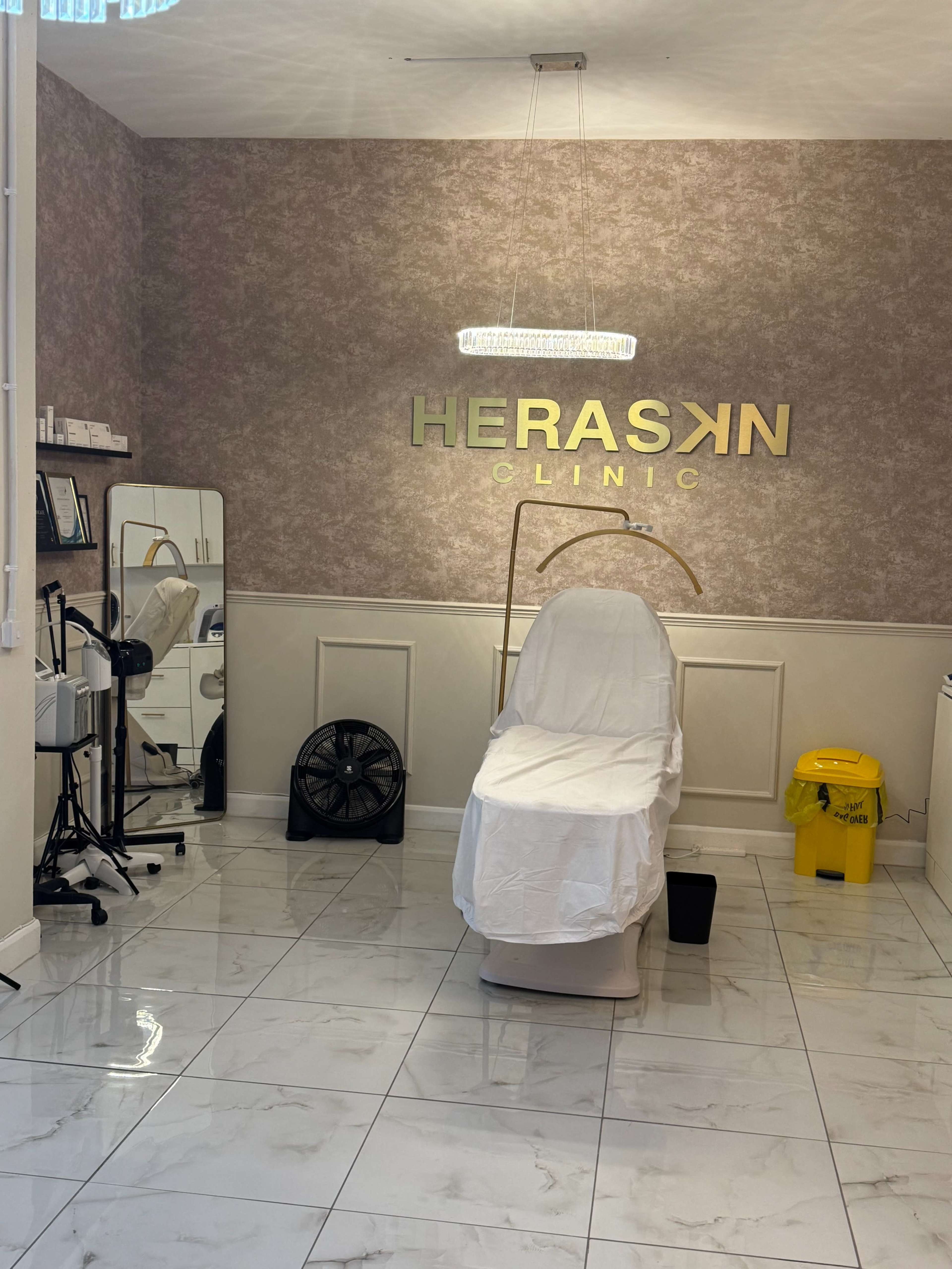 The image shows a room in a clinic featuring a treatment chair covered with a white sheet, a large mirror, and a waste bin, all against a textured wall with the name "HERASKN CLINIC" prominently displayed.