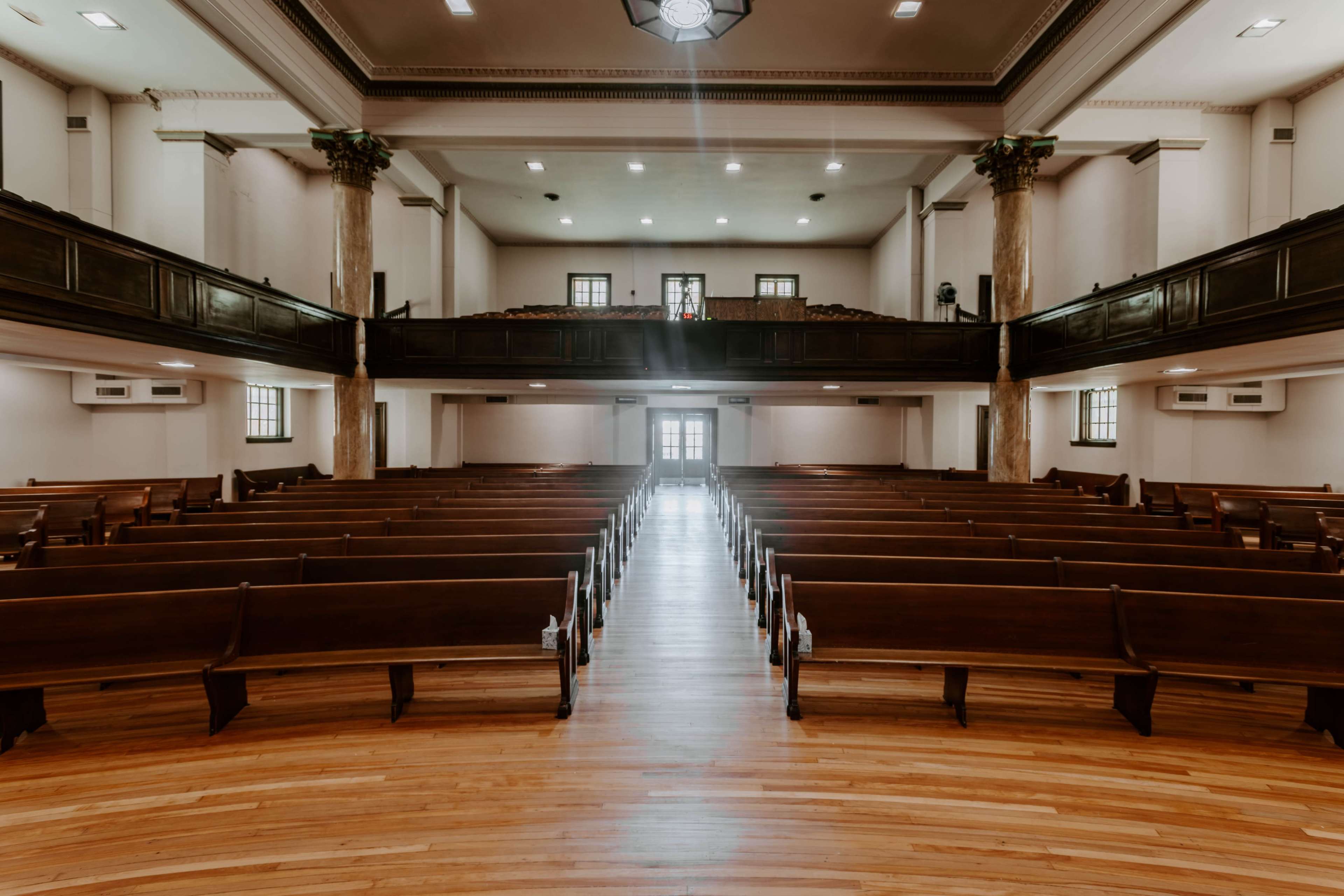 The image shows the interior of a church with rows of wooden pews facing a central aisle leading to a set of doors at the back.