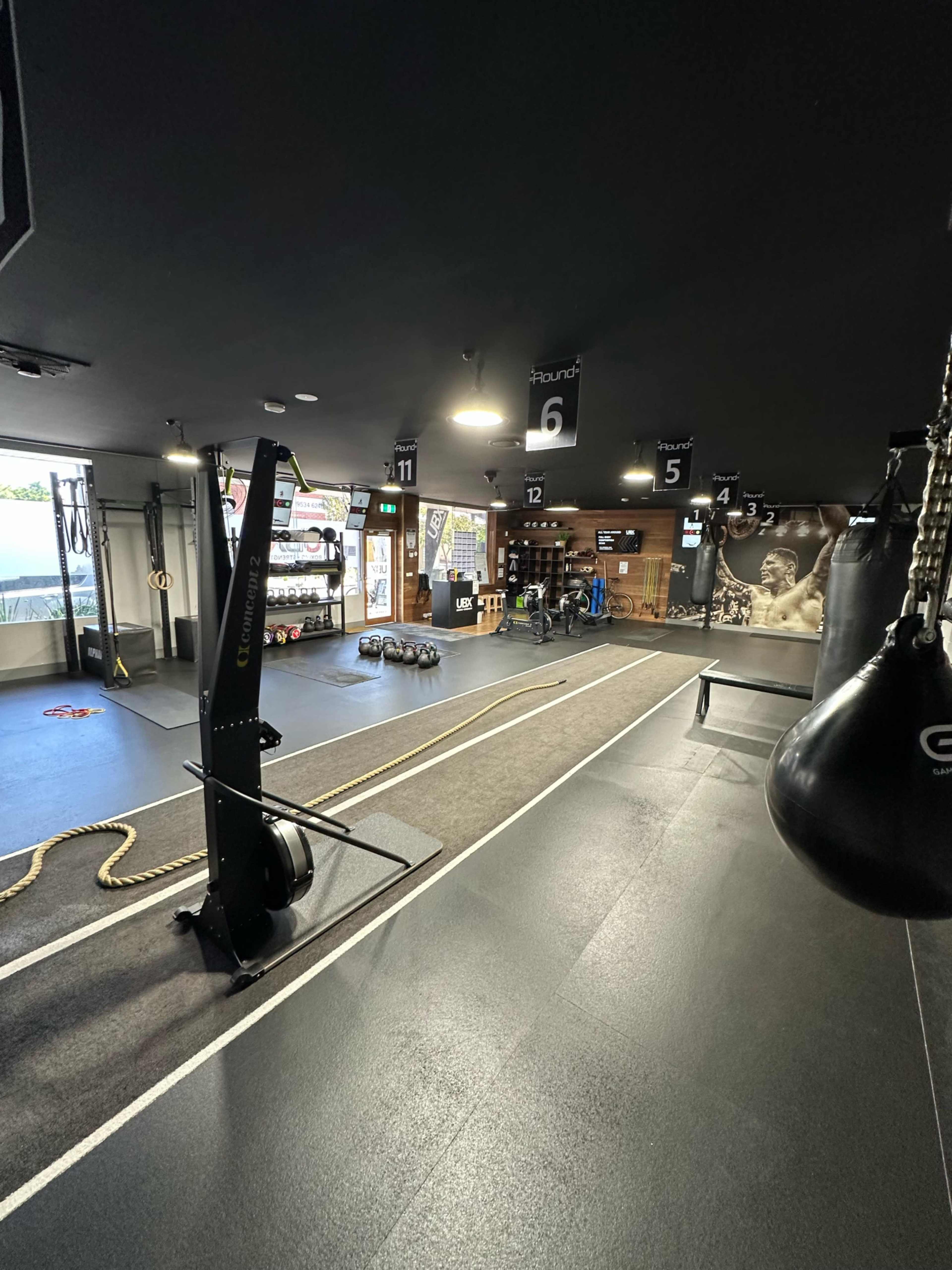 The image shows a modern gym interior with various workout equipment, including punching bags, a rope, and a space for exercise.