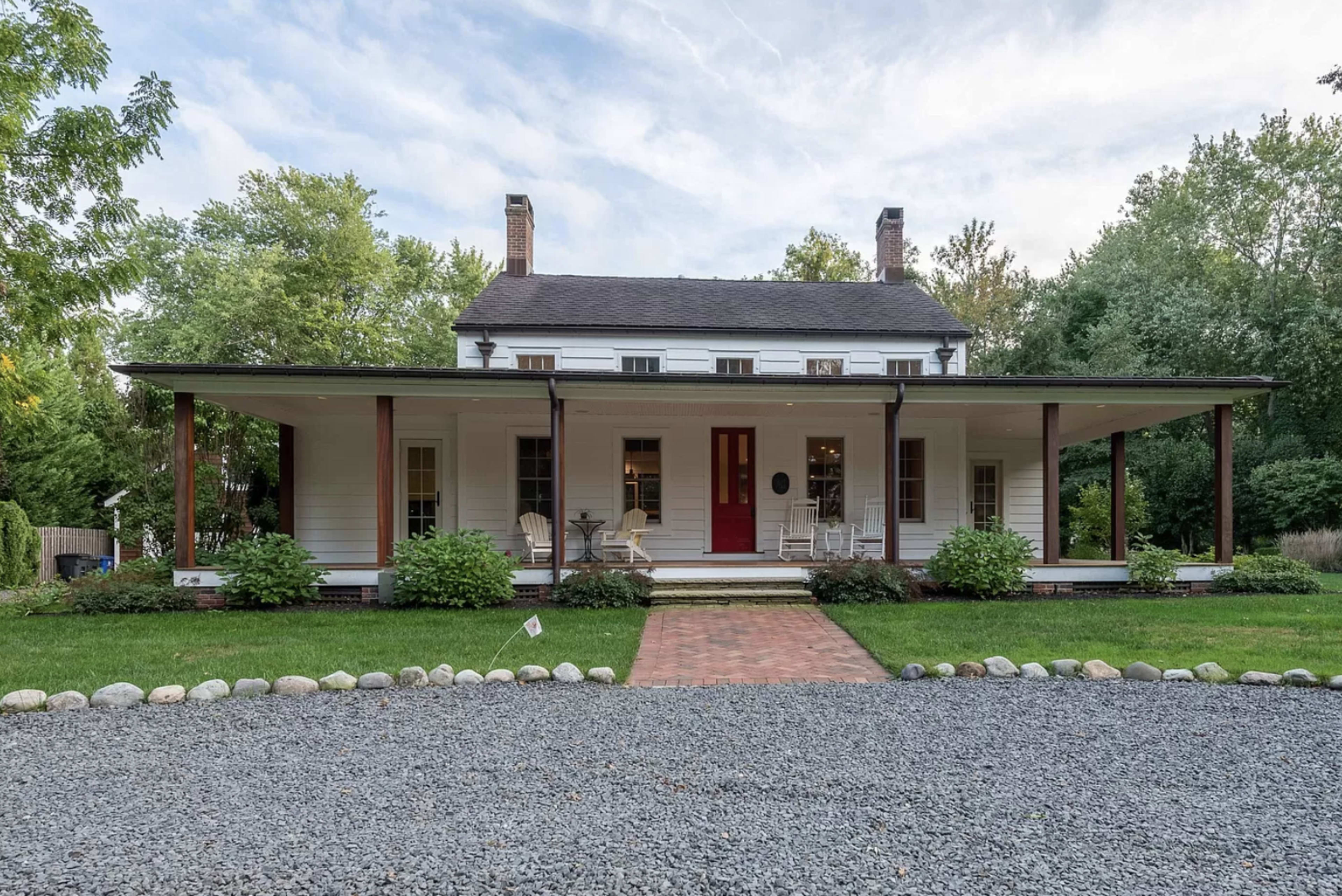 The image shows a two-story, white wooden house with a red door, surrounded by a green lawn, gravel driveway, and landscaped bushes.