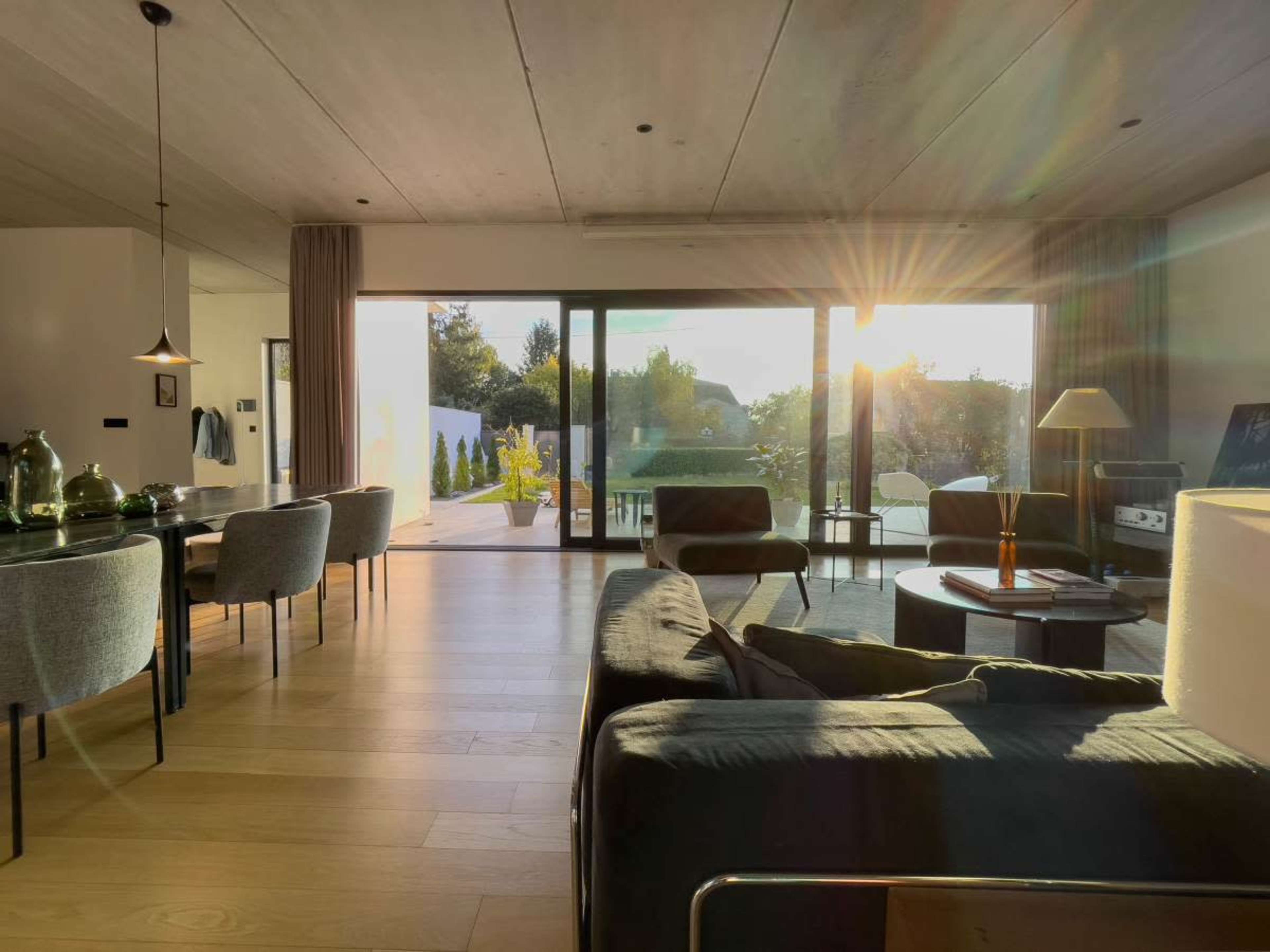 The image shows a modern living room with large glass doors opening to an outdoor space, illuminated by the warm glow of sunset.