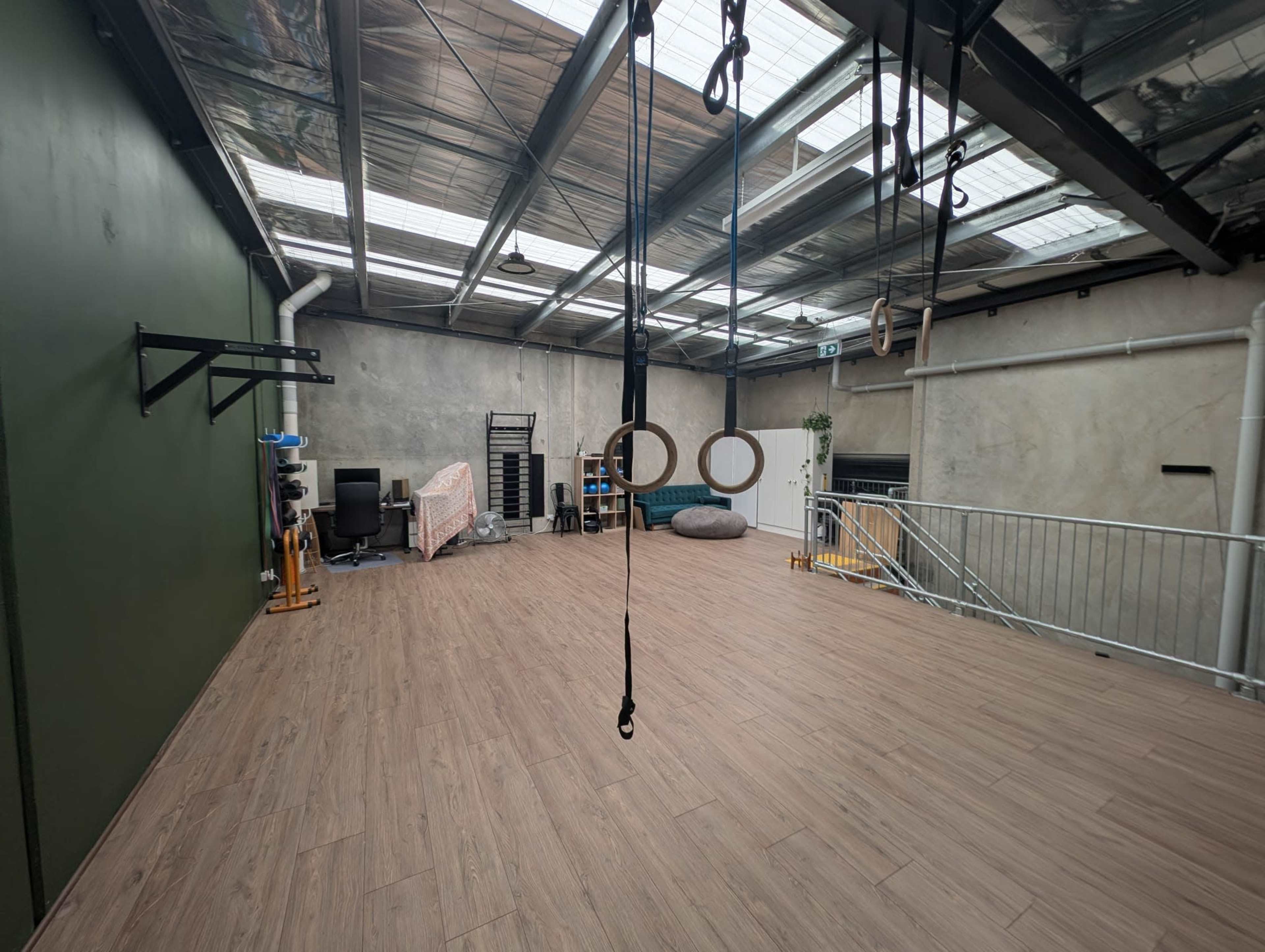 The image shows an open workout space with wooden flooring, exercise rings hanging from the ceiling, and various gym equipment, including a chair and a small couch in the background.