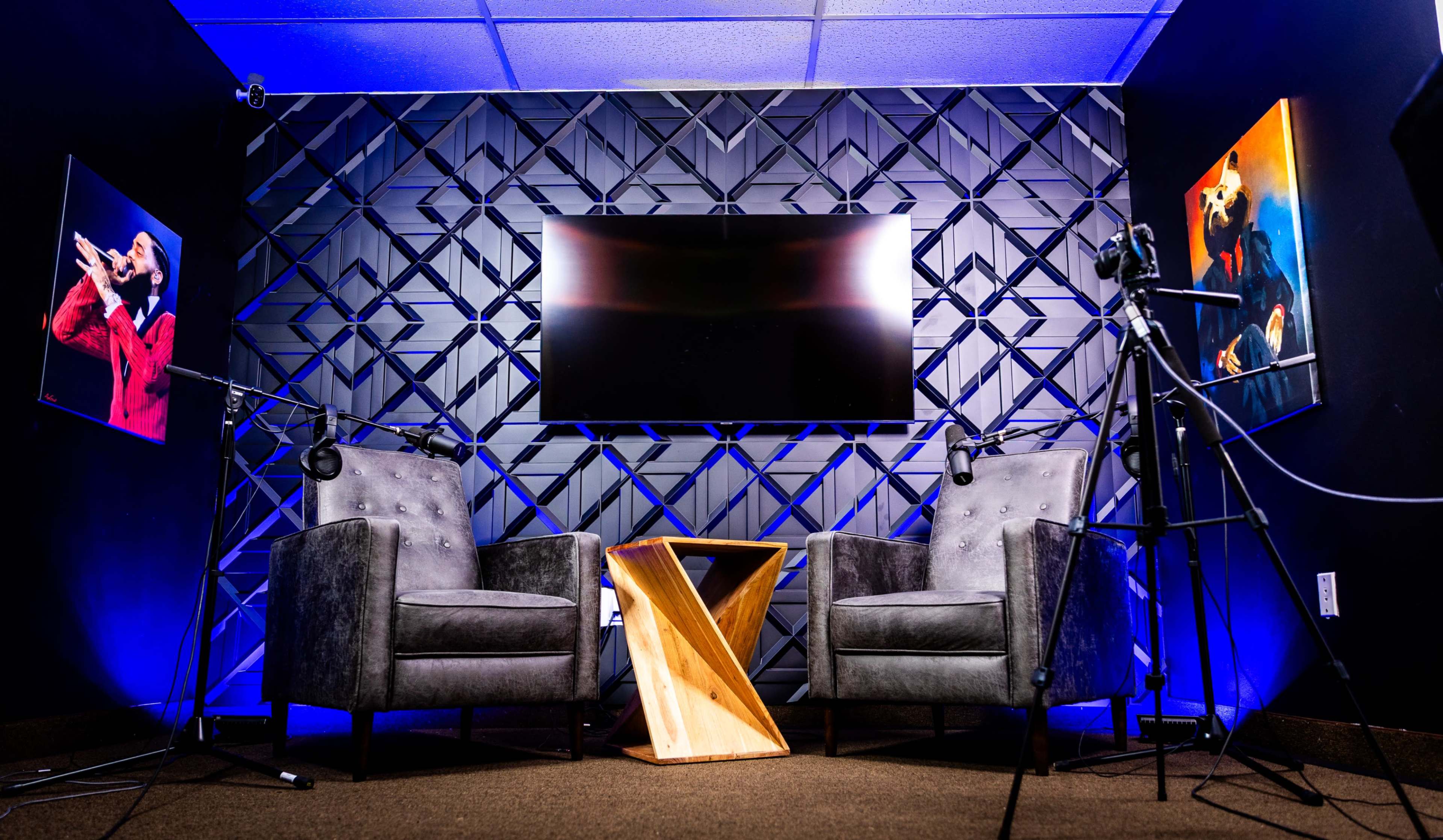 The image shows a recording studio with two gray armchairs facing a wooden table, a television mounted on a textured black wall, and artwork featuring musicians on either side.