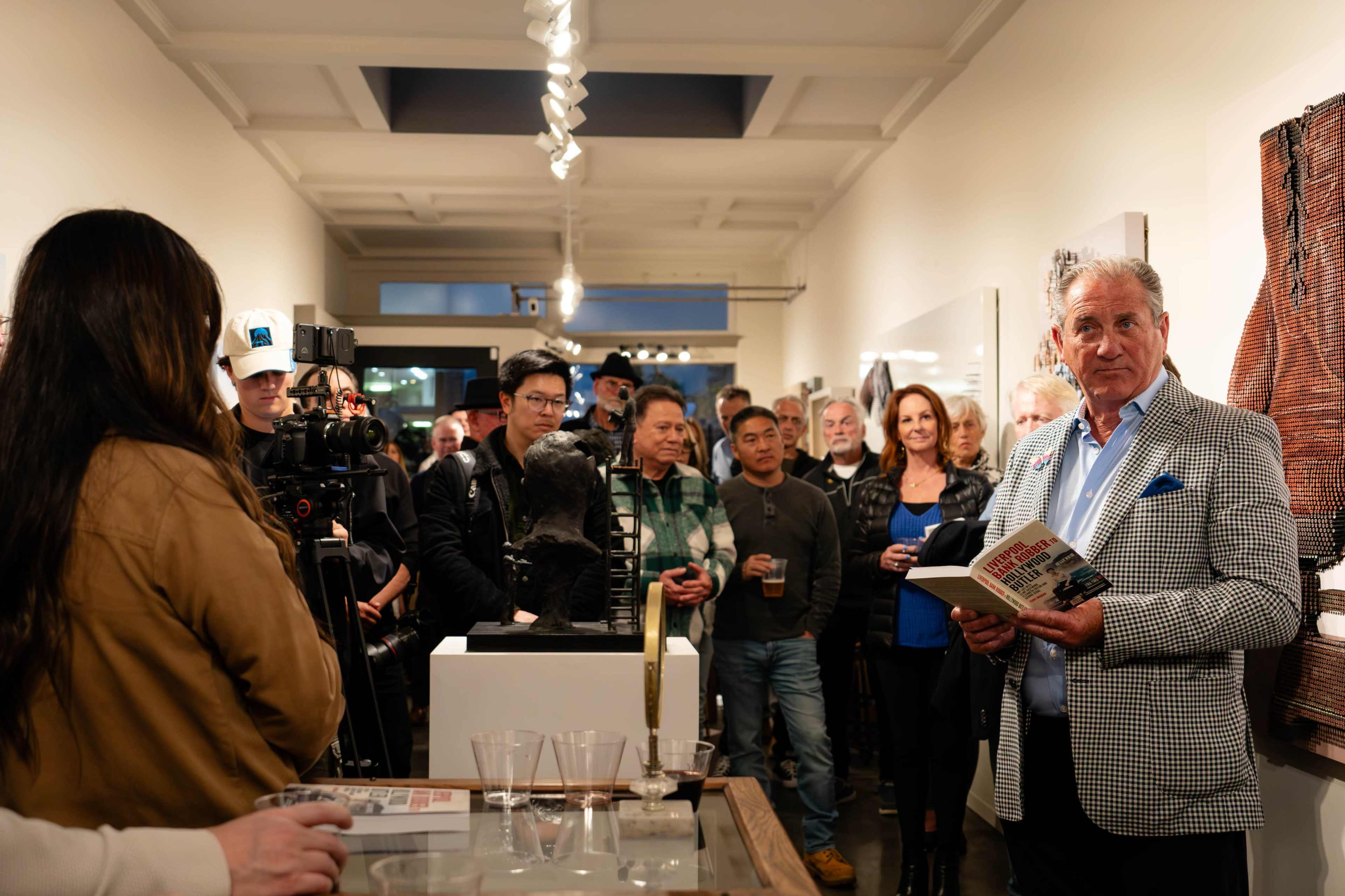 A group of people gathers in an art gallery as a man in a checked blazer reads from a book, with various artworks displayed around the room.