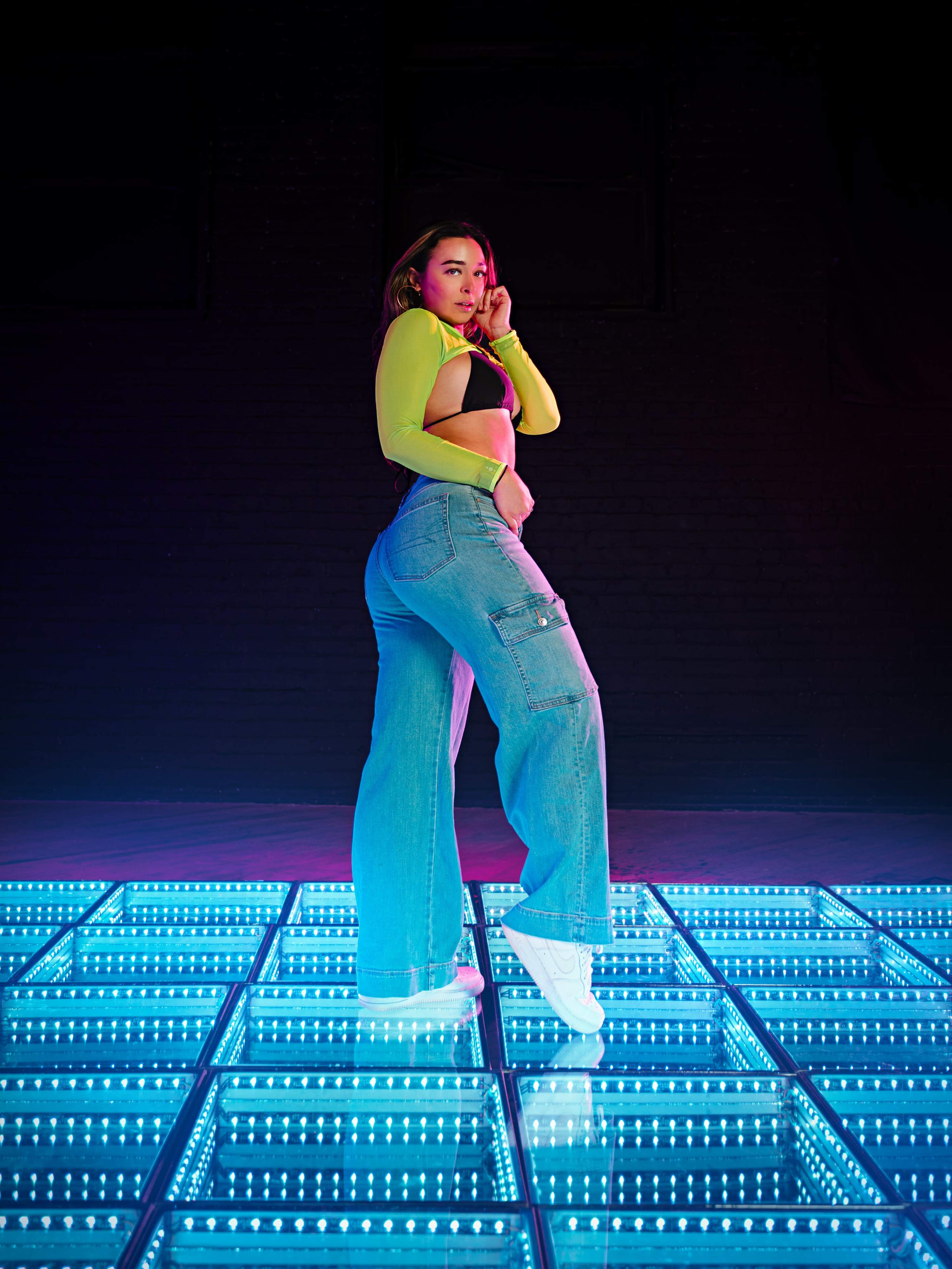 A woman poses confidently on a lighted floor in a dance studio, wearing a bright yellow crop top and baggy jeans.