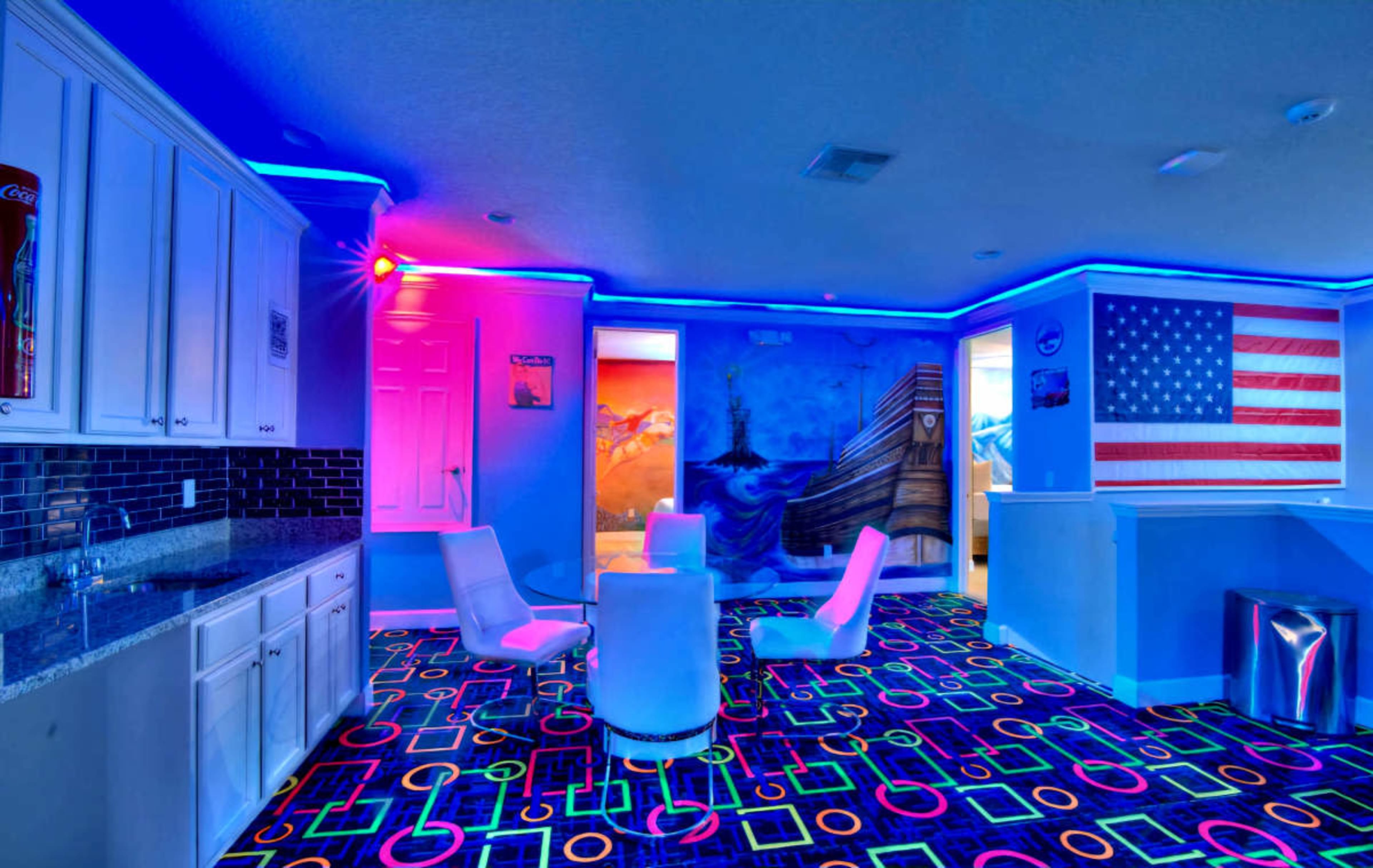 The image shows a brightly colored recreational room with neon lighting, a patterned floor, a kitchenette area, and mural artwork on the walls.