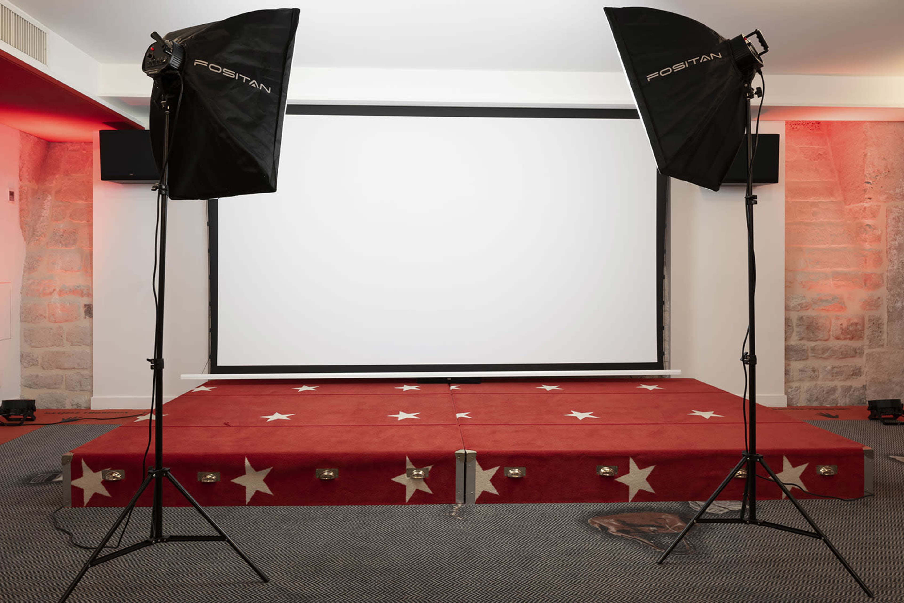A stage with a red carpet featuring stars, flanked by two lighting setups, in front of a blank projection screen.