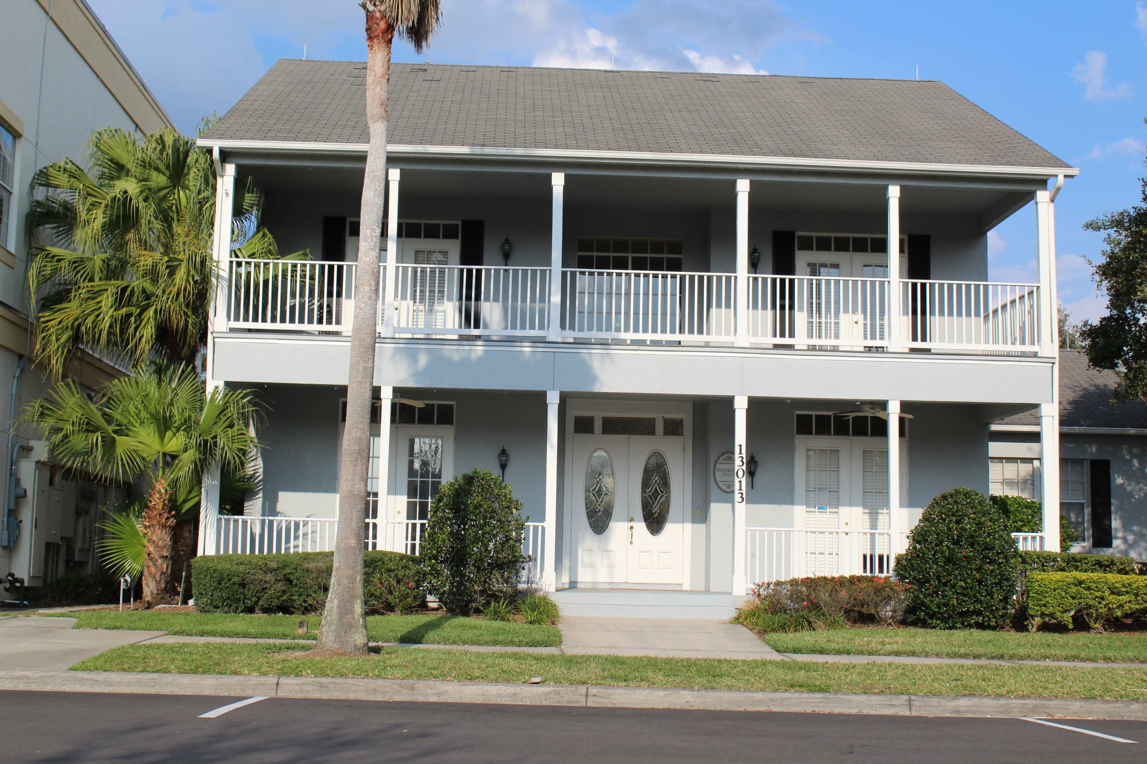 A two-story blue house with white railings and double front doors, flanked by landscaping and palm trees.