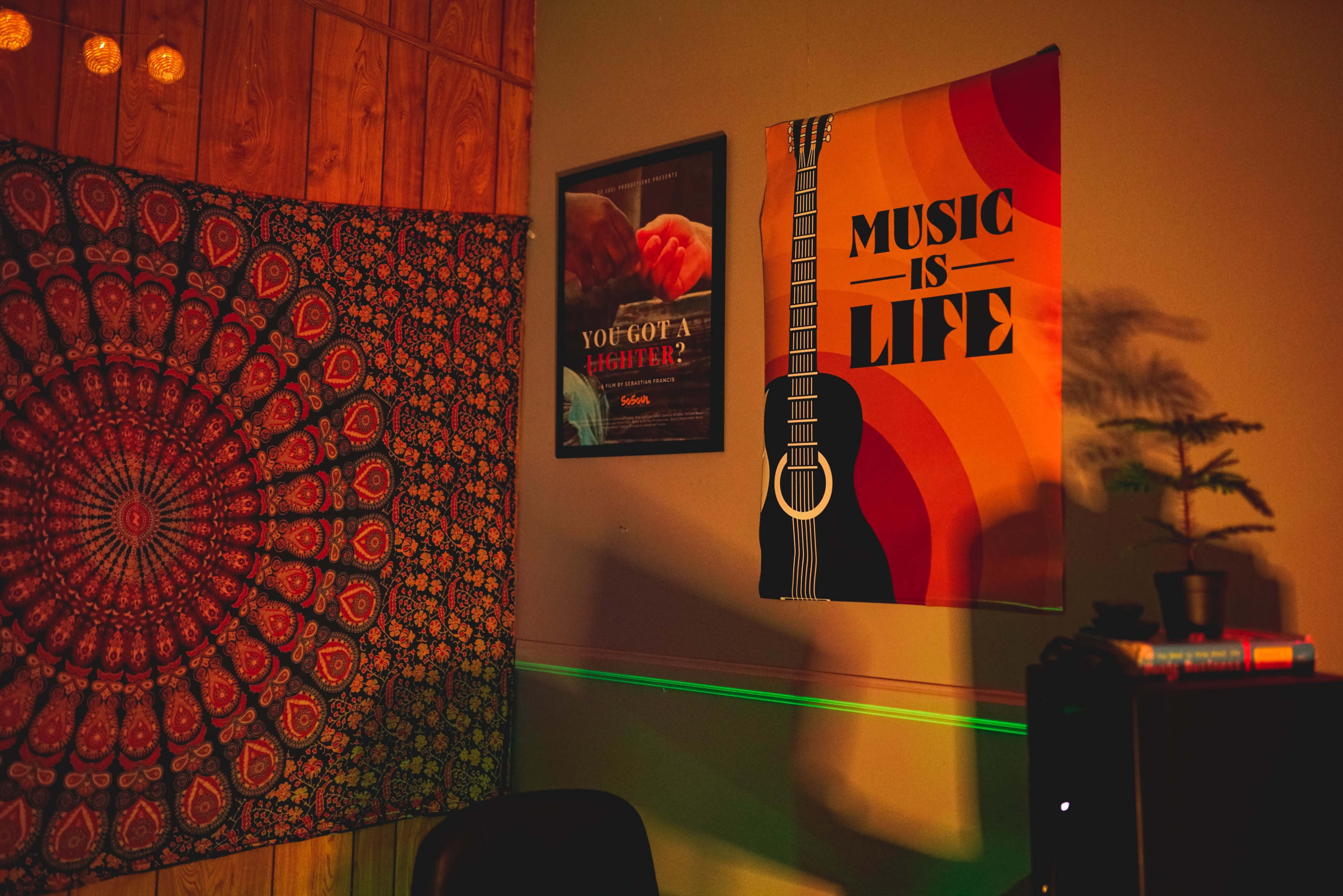 The image shows a cozy indoor room featuring a patterned tapestry on the left, a poster with a guitar and text on the right, and a plant on a shelf.