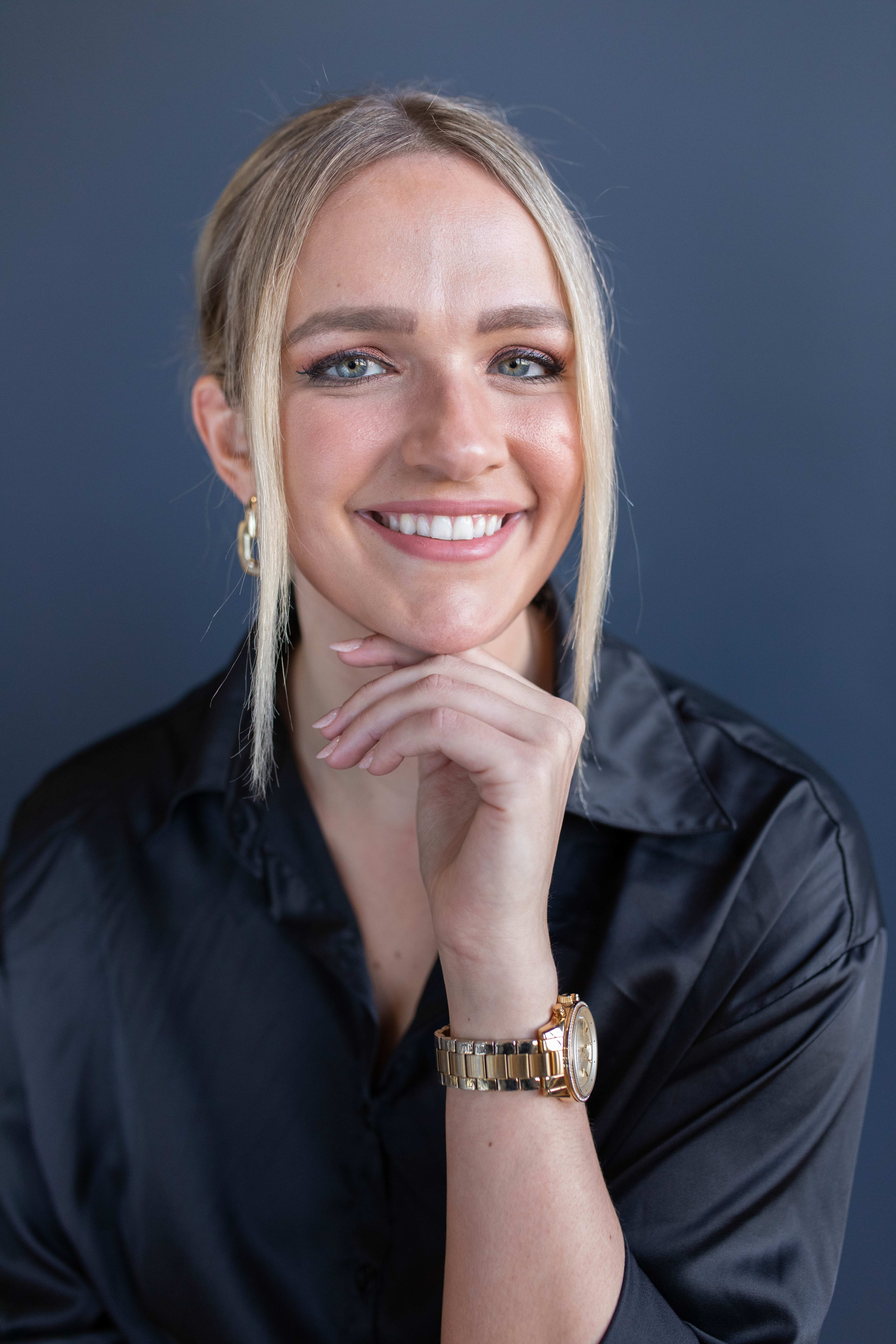 A woman wearing a black shirt and a gold watch in a head shot.