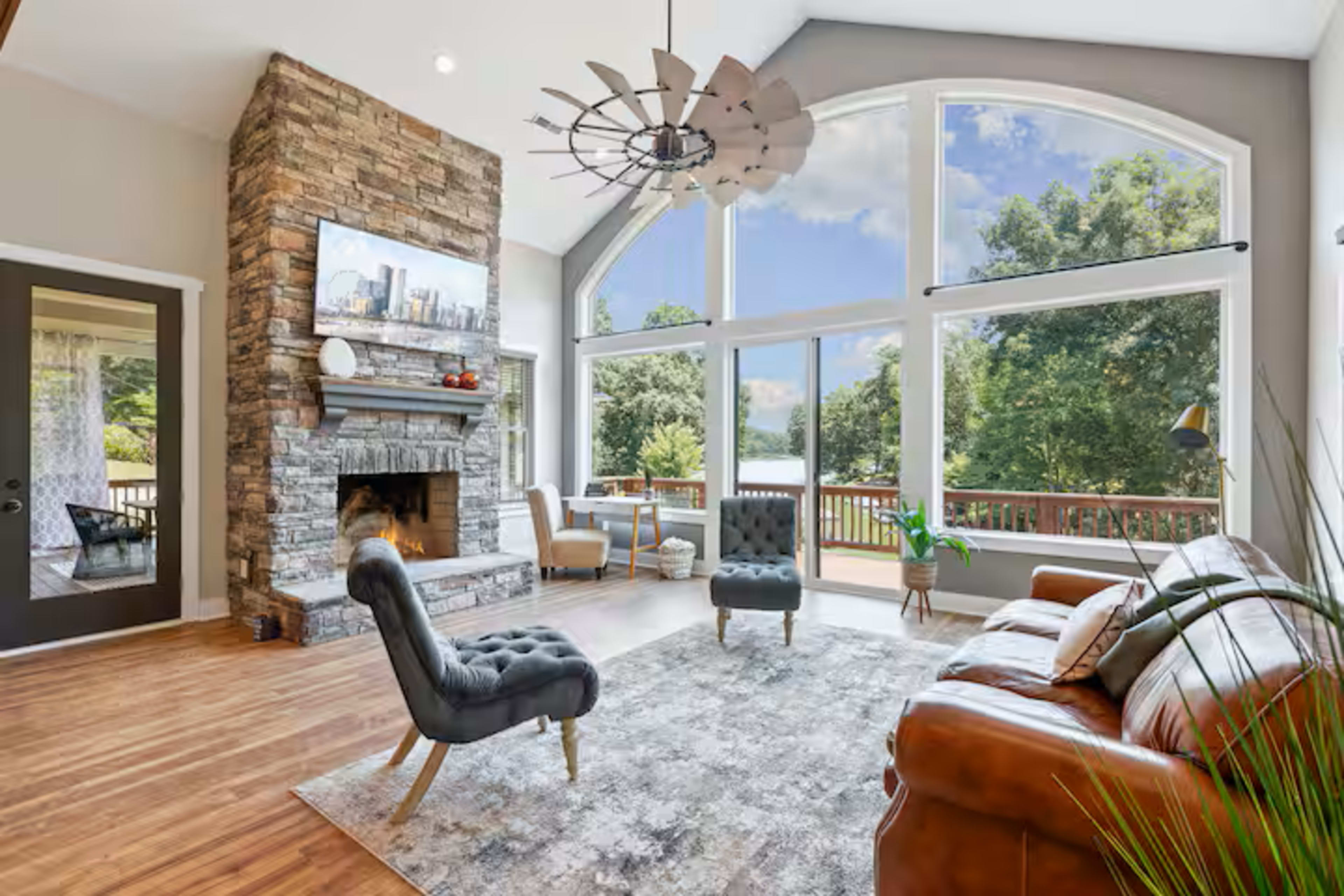 The image shows a living room featuring a stone fireplace, large windows with a view, modern furniture, and a wooden floor.
