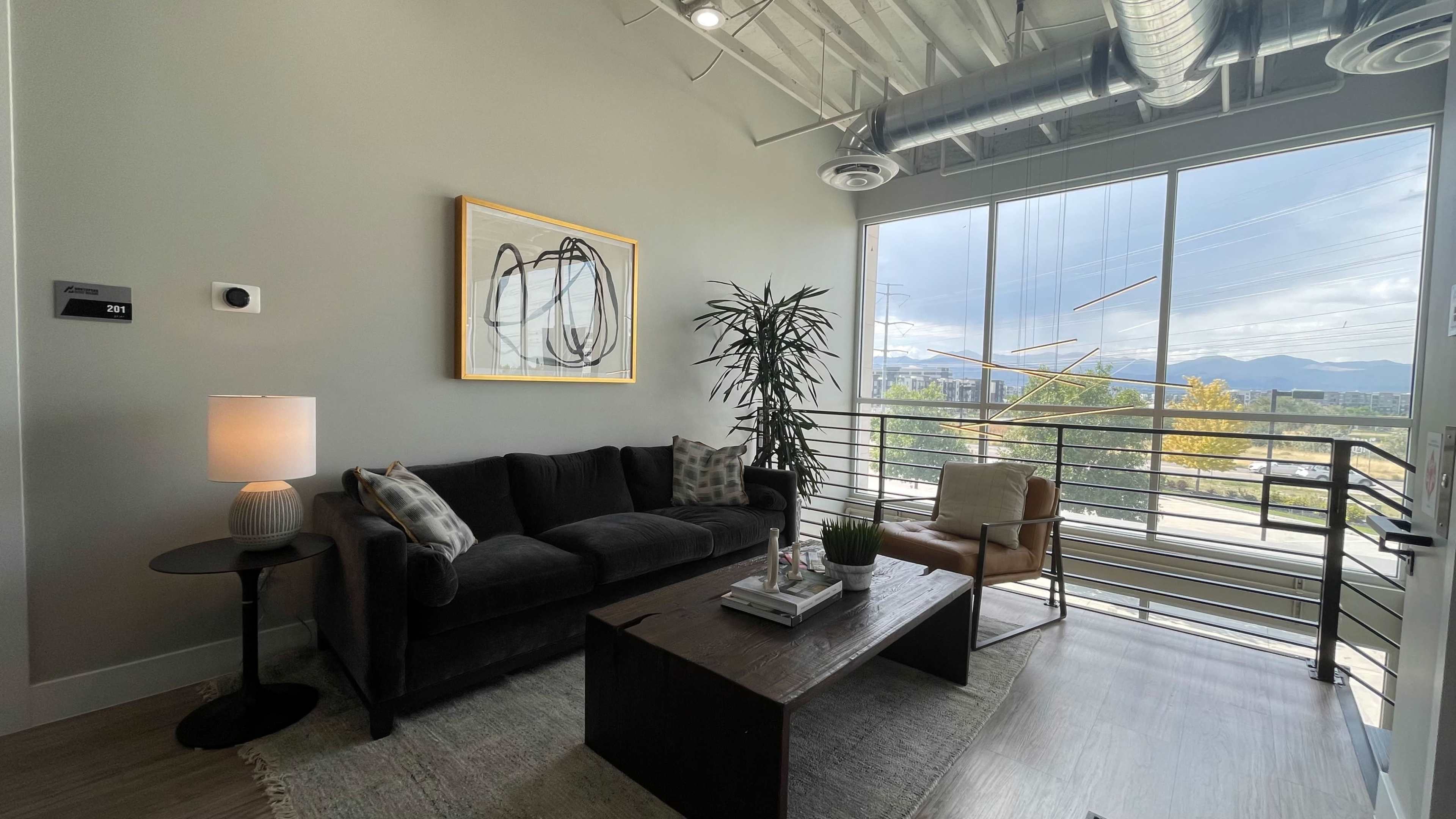A modern living room with a black sofa, a wooden coffee table, a floor lamp, and large windows overlooking a scenic view.